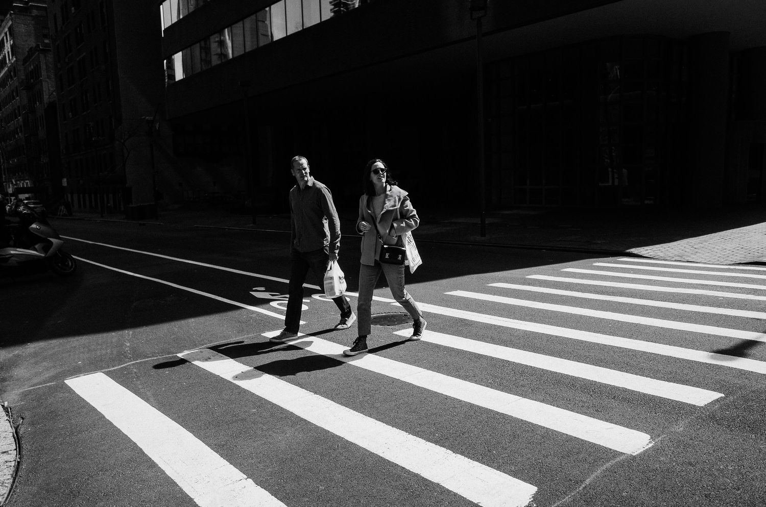 Two pedestrians cross a zebra crossing in dramatic black and white lighting on a sunny urban street.