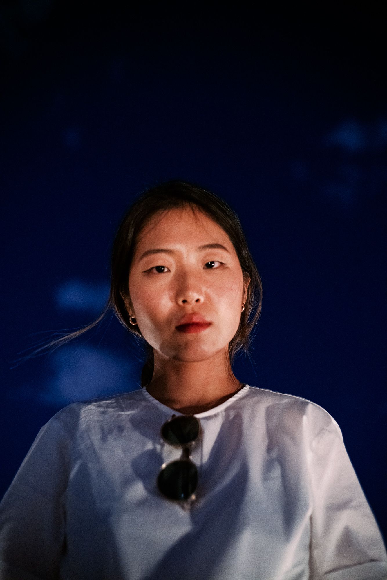 Portrait of young woman in white shirt against deep blue background with sunglasses on collar.