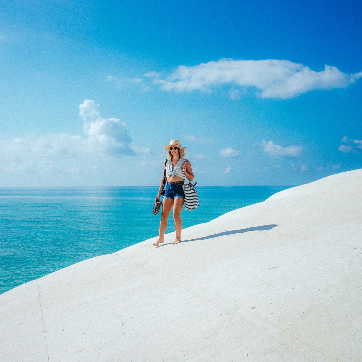 Person in a white dress walking along pristine white sand beach with turquoise ocean waters and blue sky in the background.