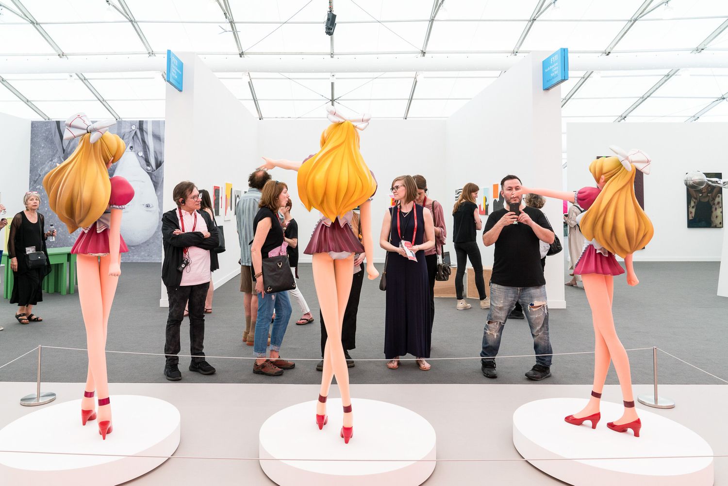 Visitors explore a contemporary art fair featuring large anime-style figurine sculptures on white pedestals in a bright gallery.