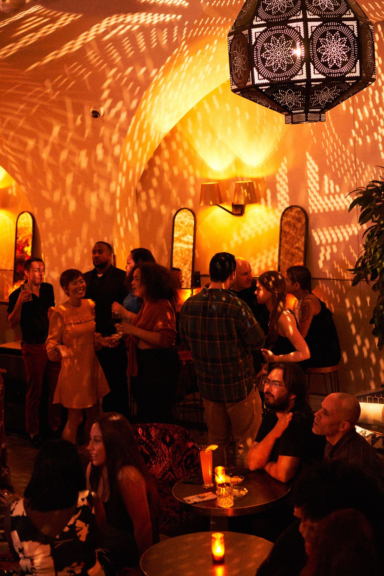 Guests mingle in a warmly lit bar with ornate lantern casting geometric shadows on arched golden walls.