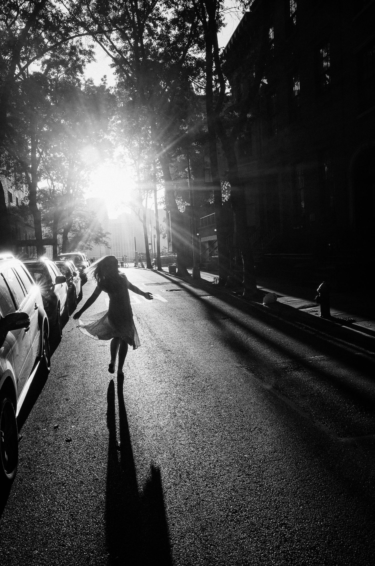 A silhouette dances in a sunlit urban street casting long shadows among parked cars in black and white photography.