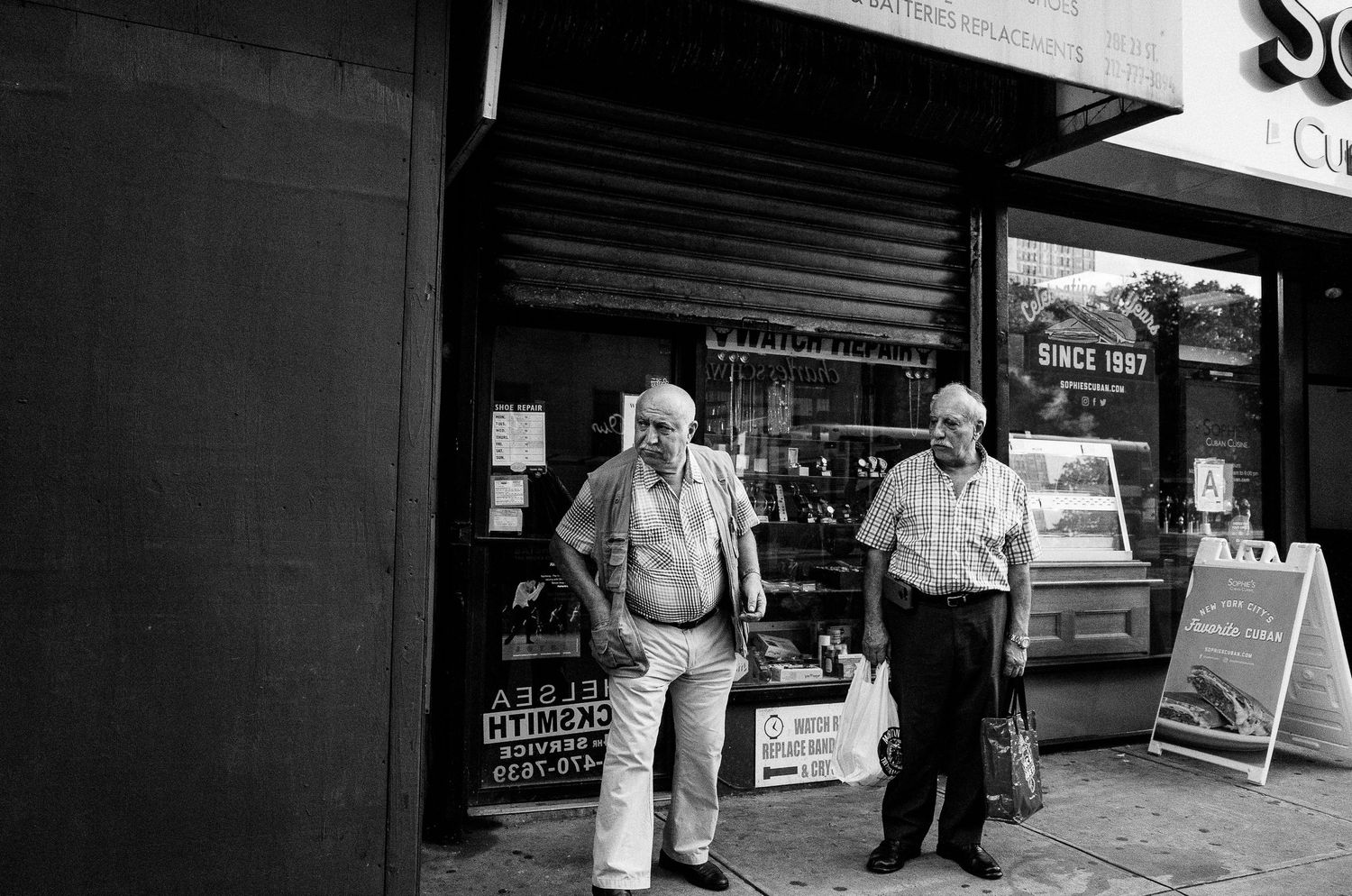 Two elderly men stand outside a storefront on a sidewalk in a black and white photograph.