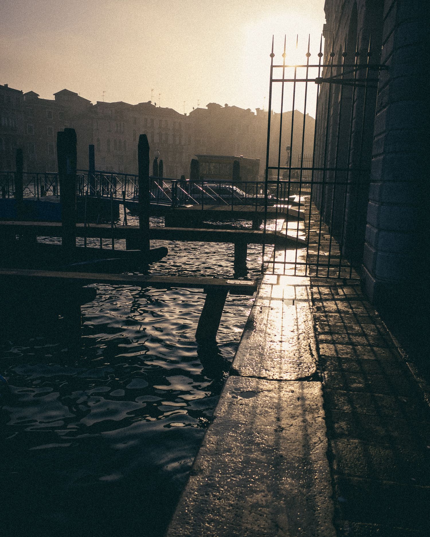 Sunlight reflects off wet cobblestone walkway along a waterfront dock during sunset creating dramatic shadows.