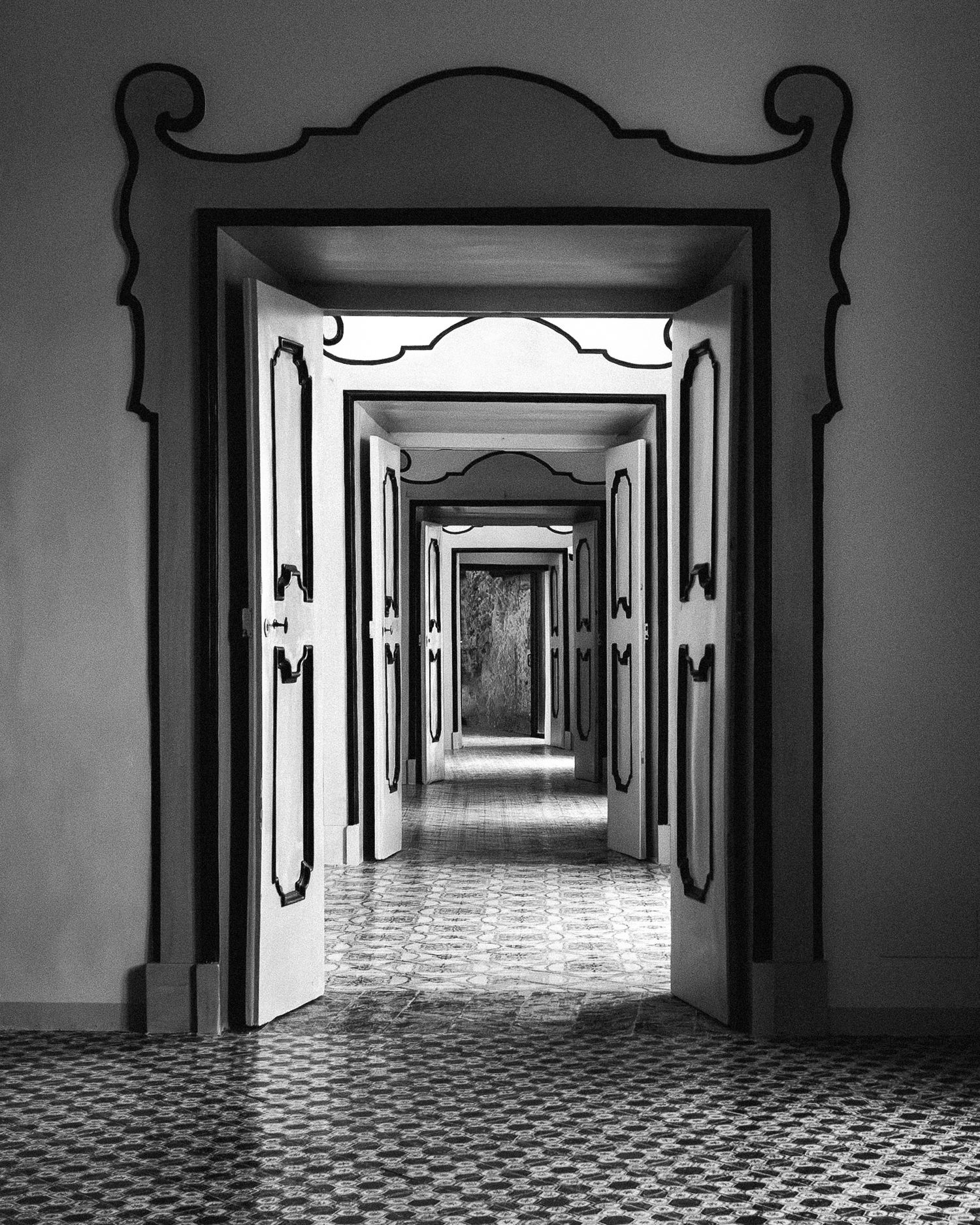 A black and white photograph shows a long empty hallway with ornate doorways and a checkered floor pattern extending into the distance.
