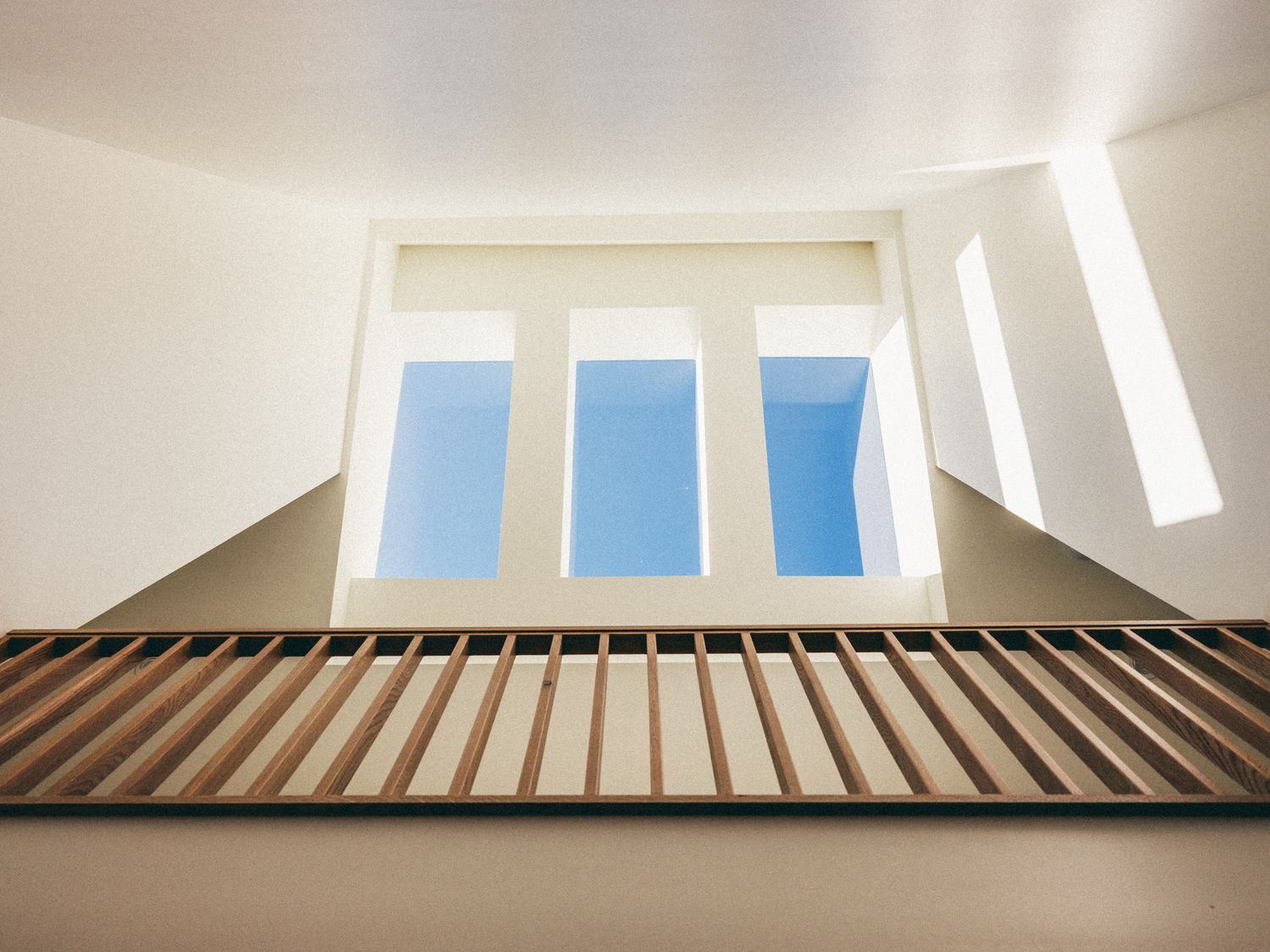Upward view of three arched windows with blue sky, white walls, and wooden slat railing in modern architecture.