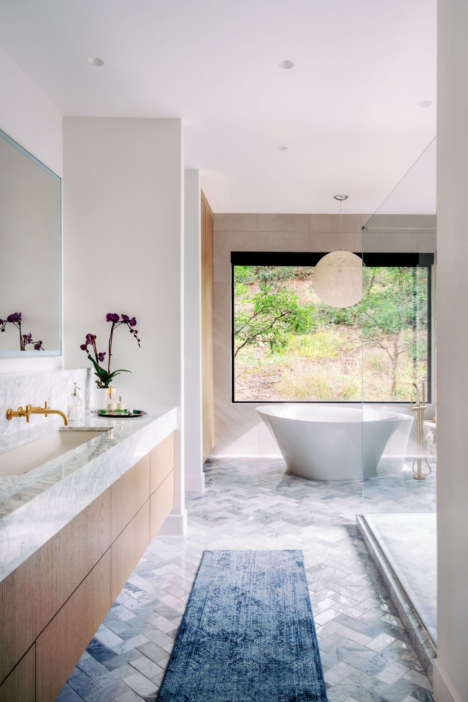 Minimalist bathroom with herringbone tile floor and wooden vanity showcases large windows overlooking nature.