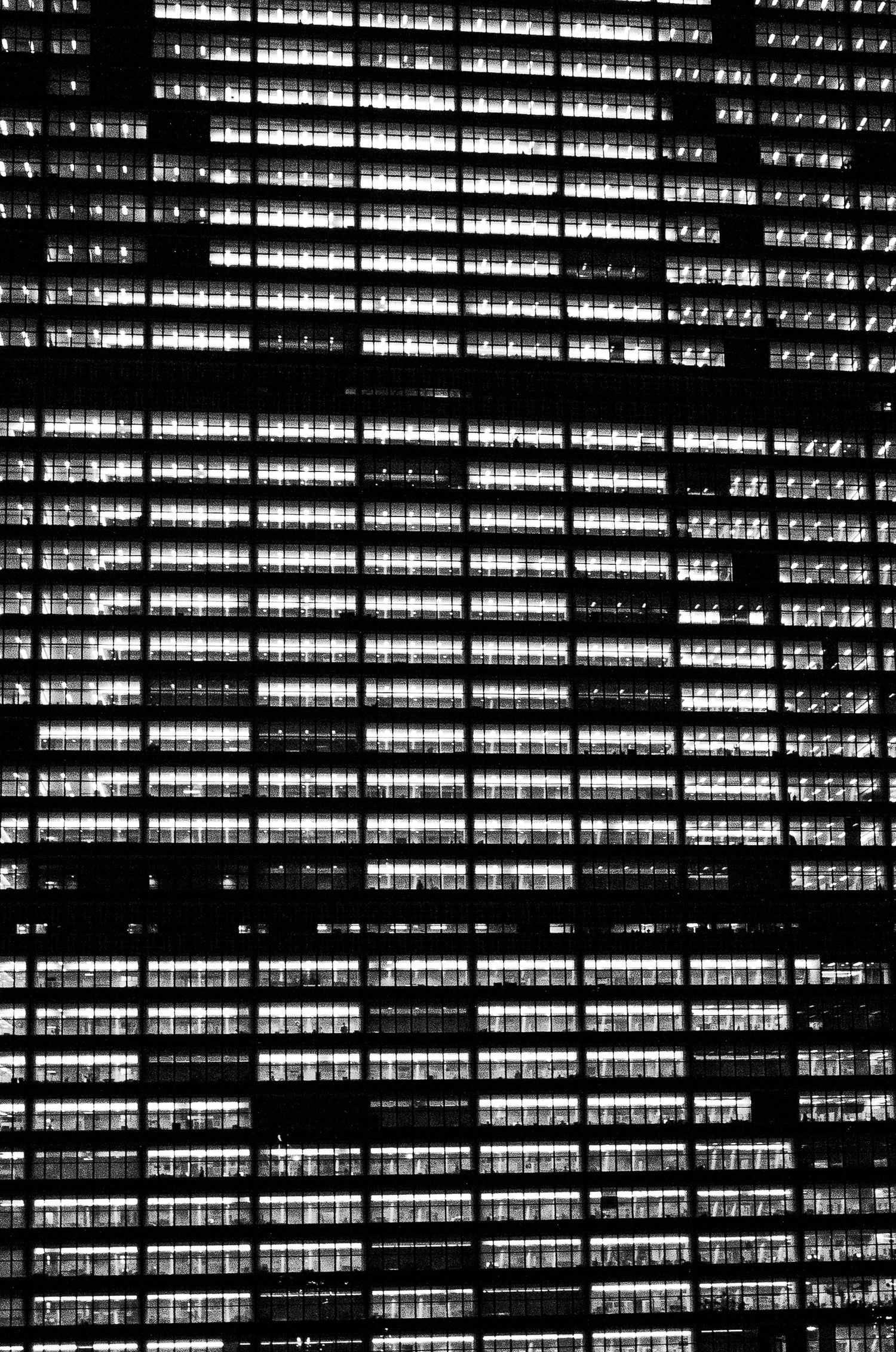 A black and white aerial view of illuminated office building windows creating a geometric pattern at night.