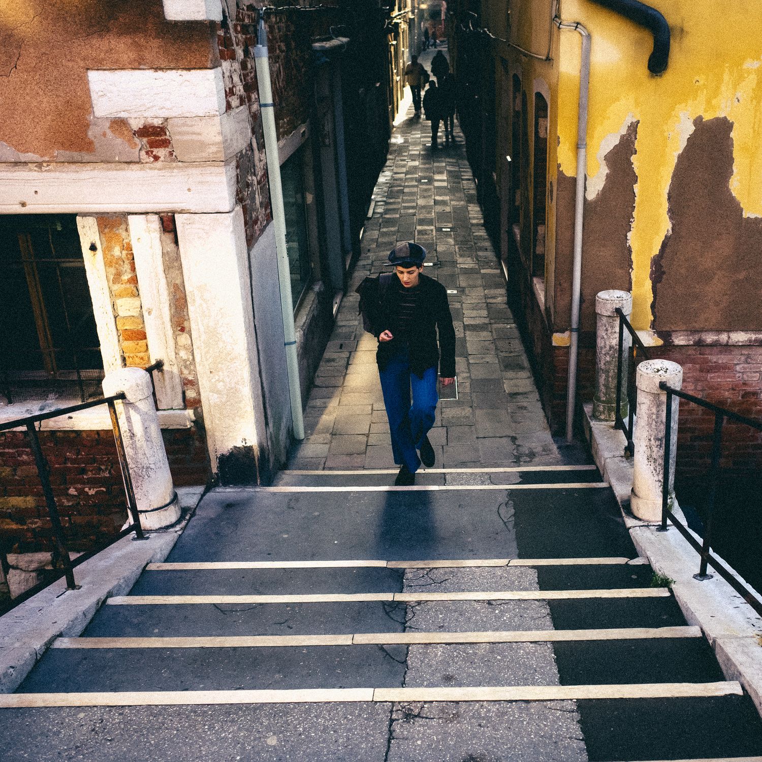 A person walks down a narrow alley with yellow walls and stone steps in an urban setting.