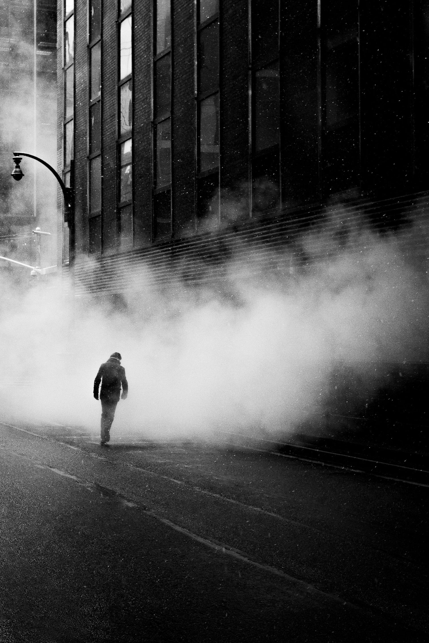 A silhouetted figure walks through atmospheric fog on a dark city street at night in a moody black and white scene.
