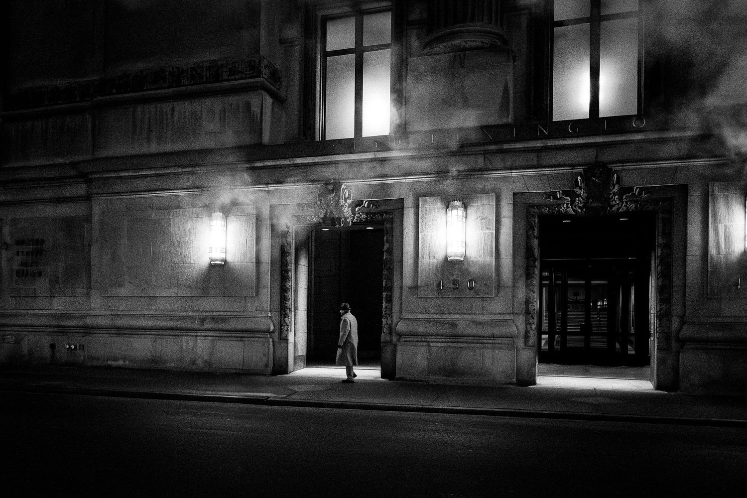 Dramatic black and white architectural photograph showing illuminated windows and wall sconces on a building at night.
