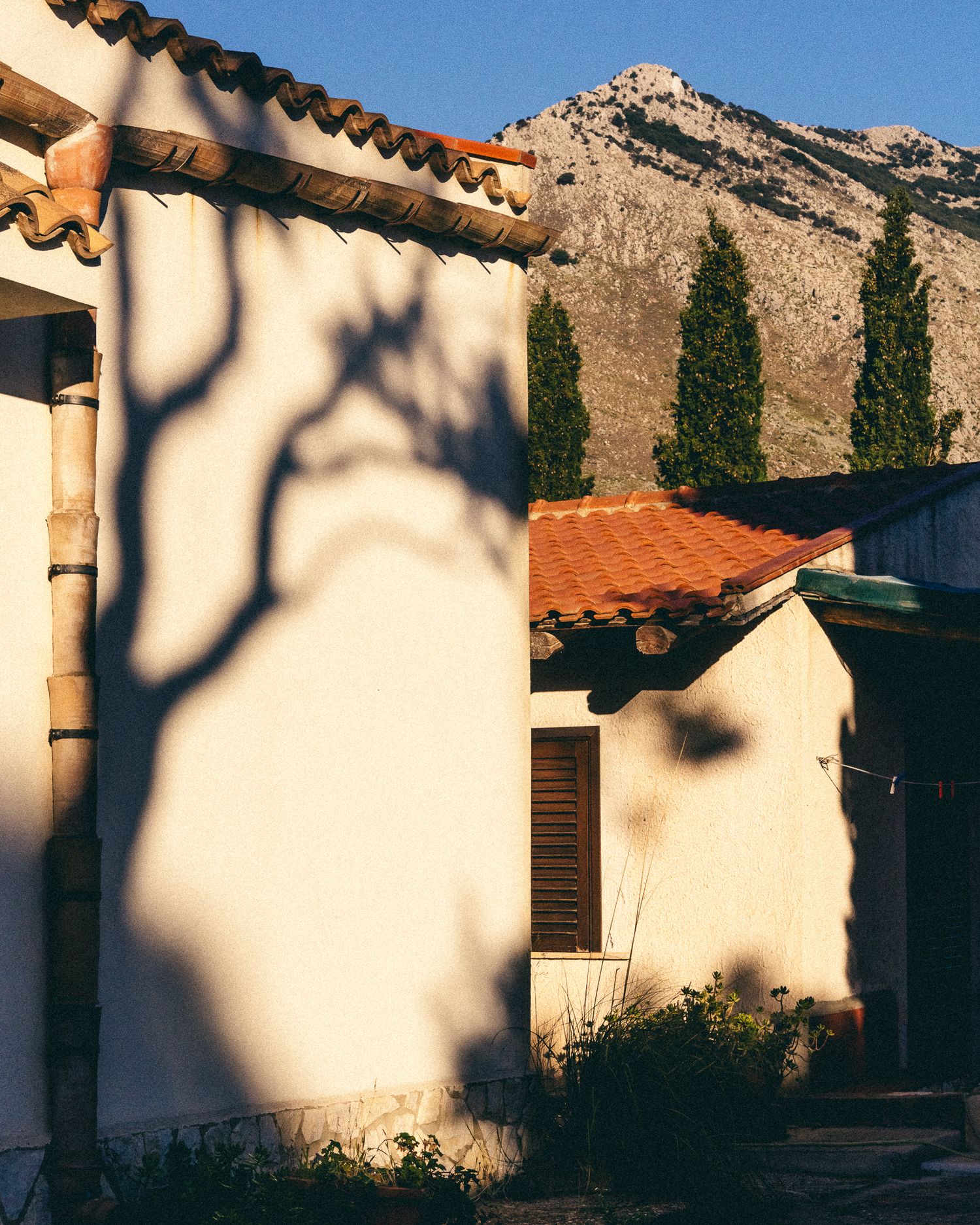 Mediterranean villa with white walls and terracotta roof against rocky mountain backdrop in golden sunlight.