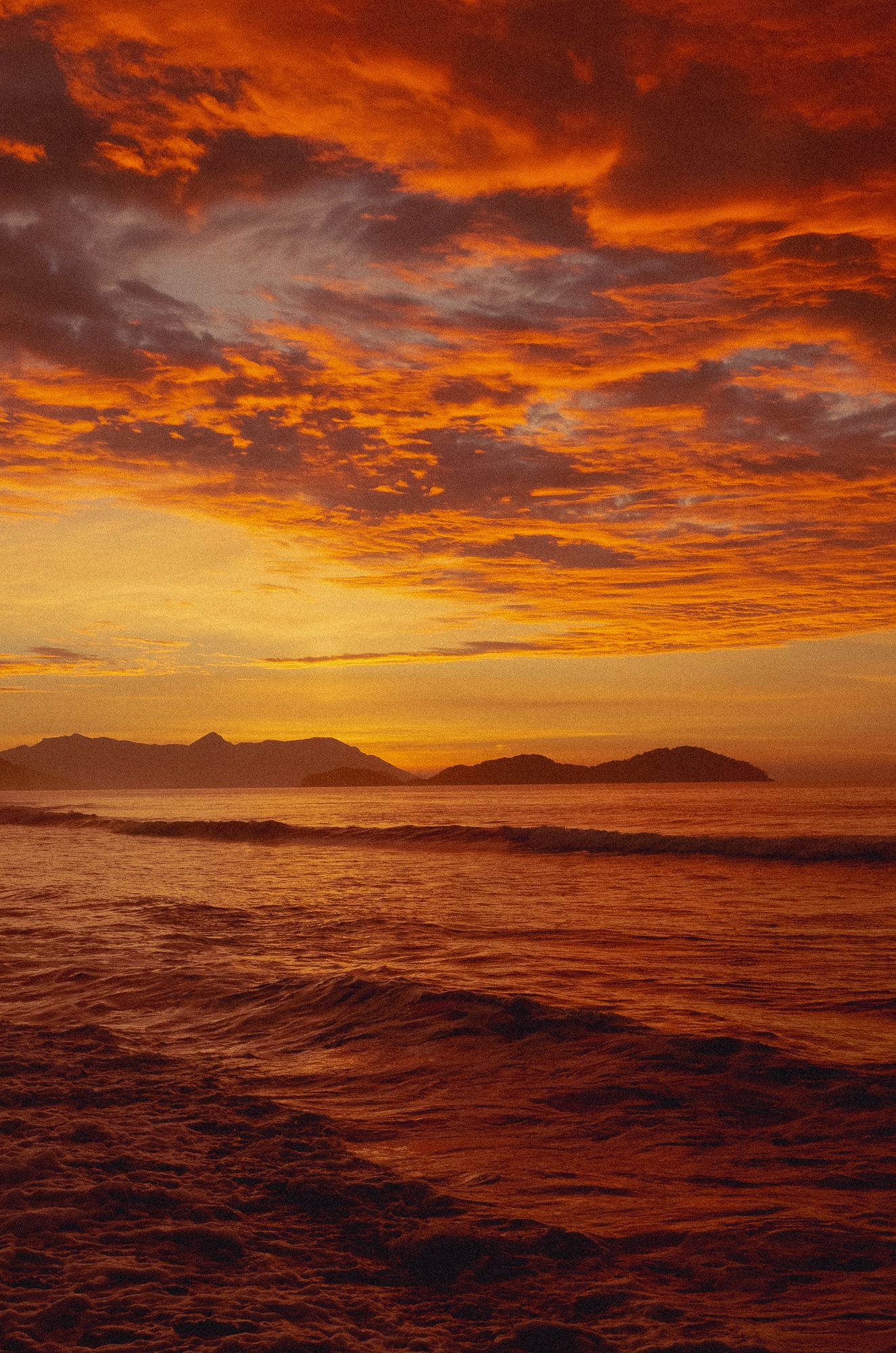 Dramatic orange and red sunset sky over ocean waves with distant mountains silhouetted on the horizon.
