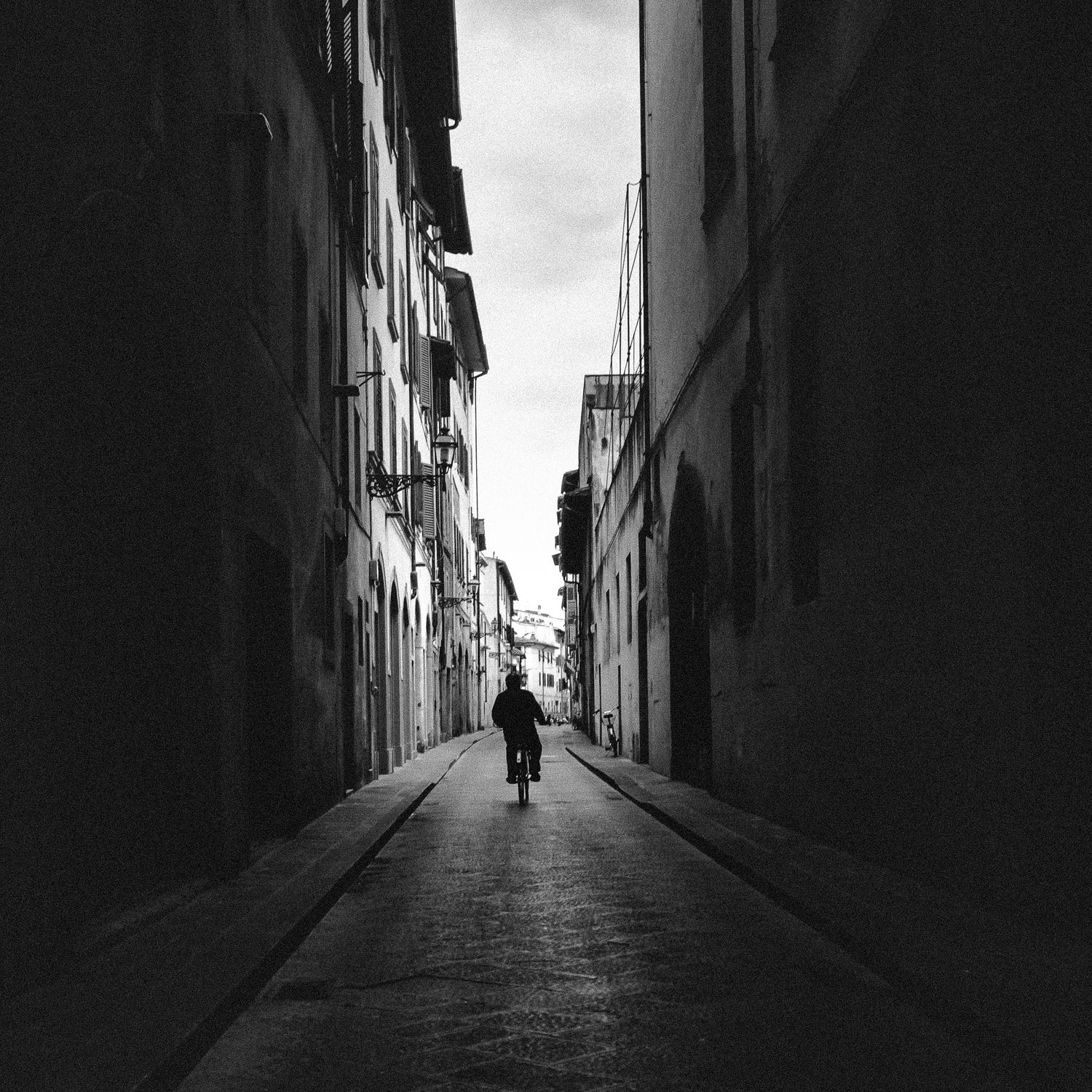 A lone figure walks down a narrow alleyway between tall buildings casting dramatic shadows in black and white.