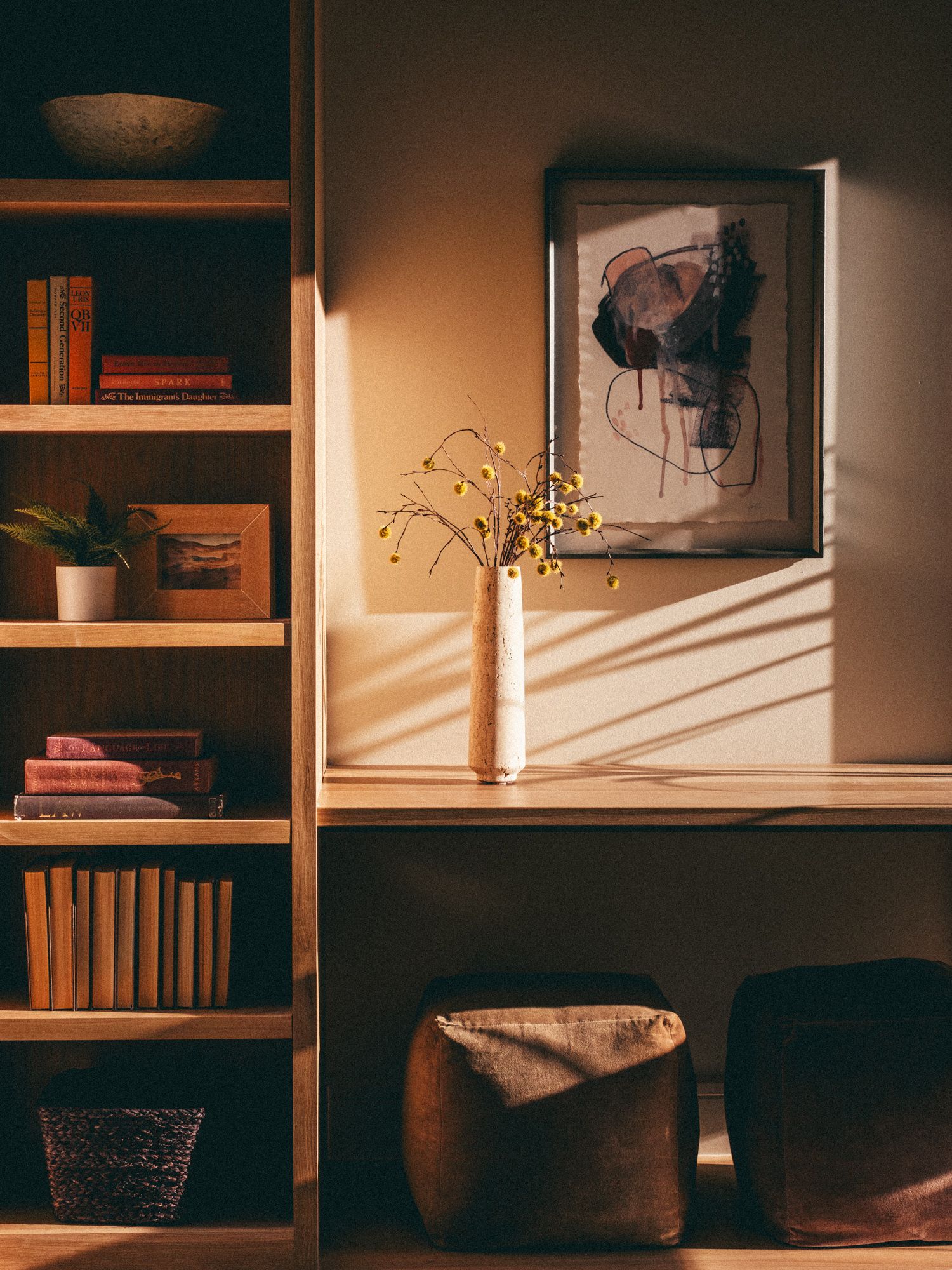 Cozy wooden bookshelf with books beside a desk featuring abstract wall art and yellow floral stems in warm sunlight.