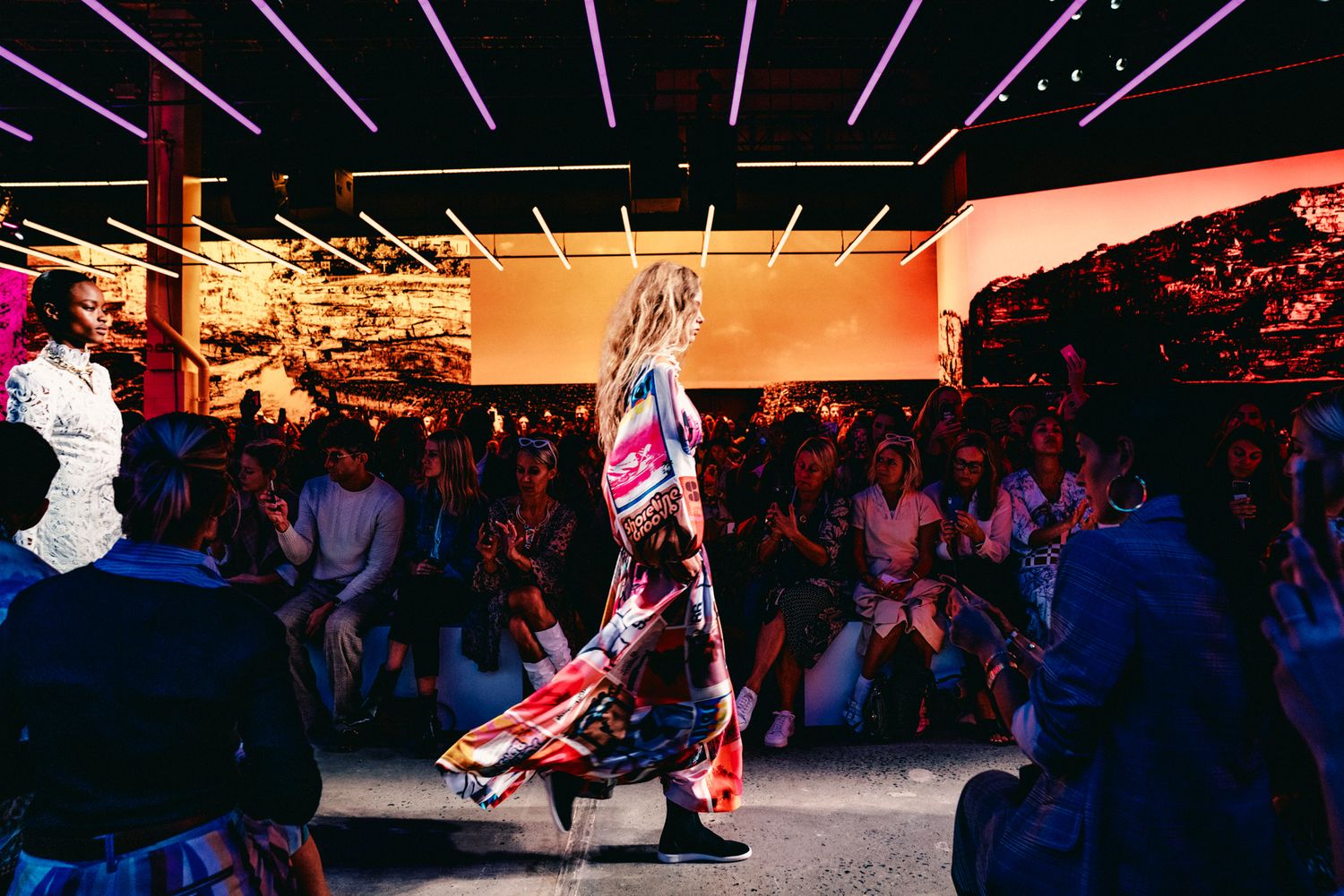 Model walks runway in colorful maxi dress at fashion show with neon lights and seated audience.
