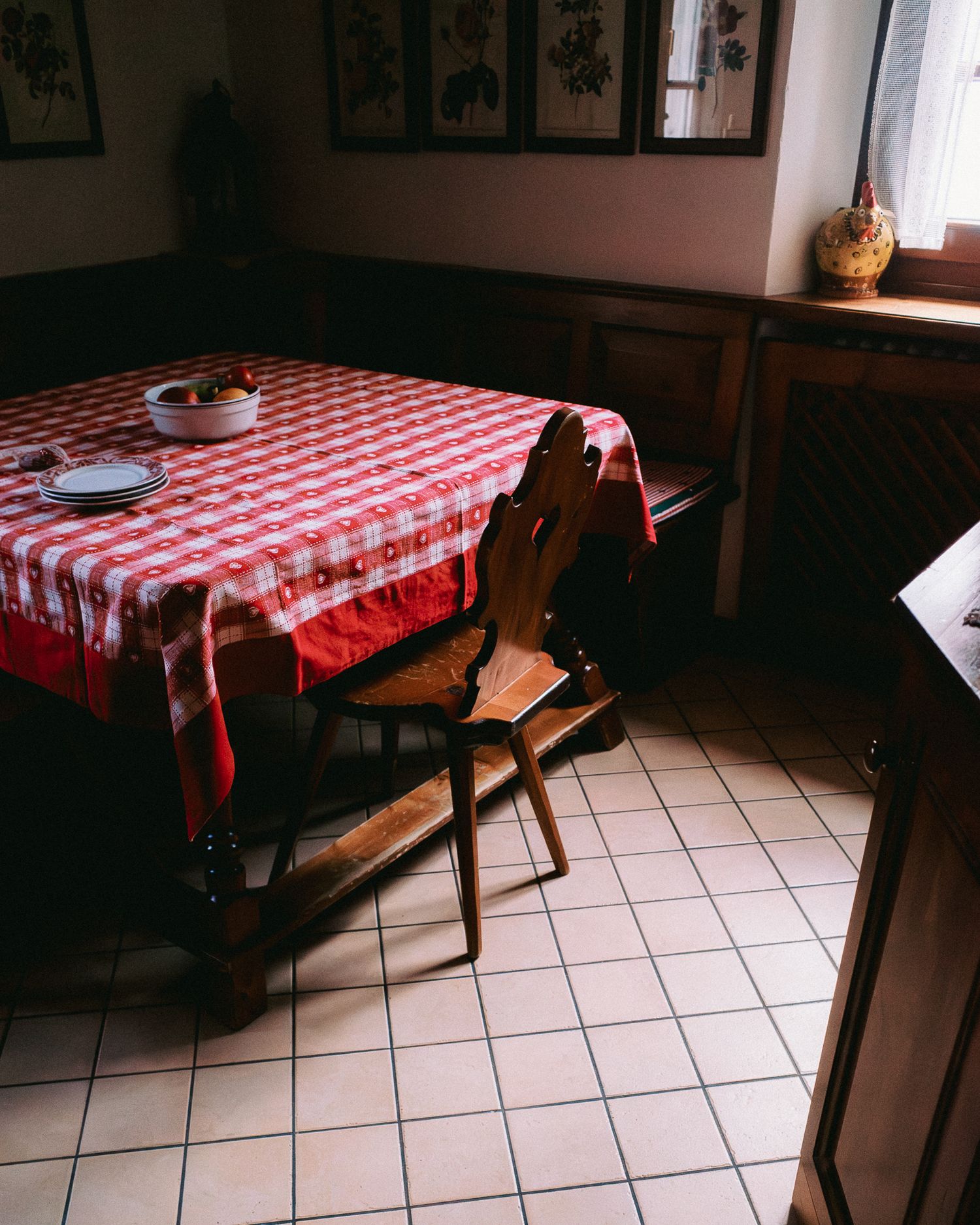 Rustic dining table with red and white checkered tablecloth and wooden chairs in a dimly lit room with tiled floor.