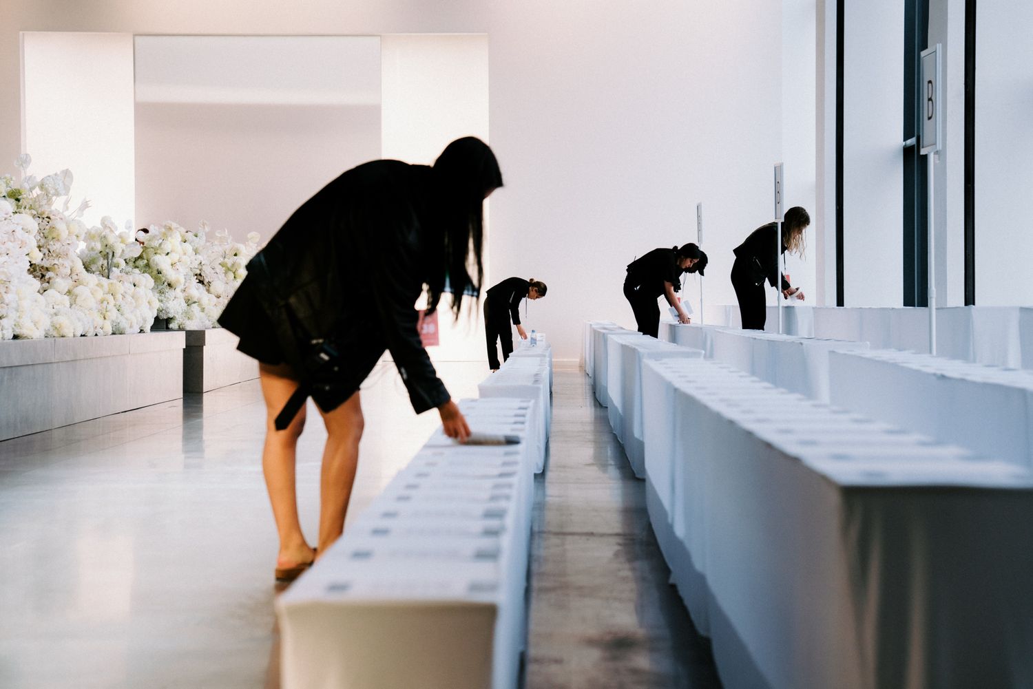 Women arranging white boxes at minimalist art installation with white flowers in bright gallery.