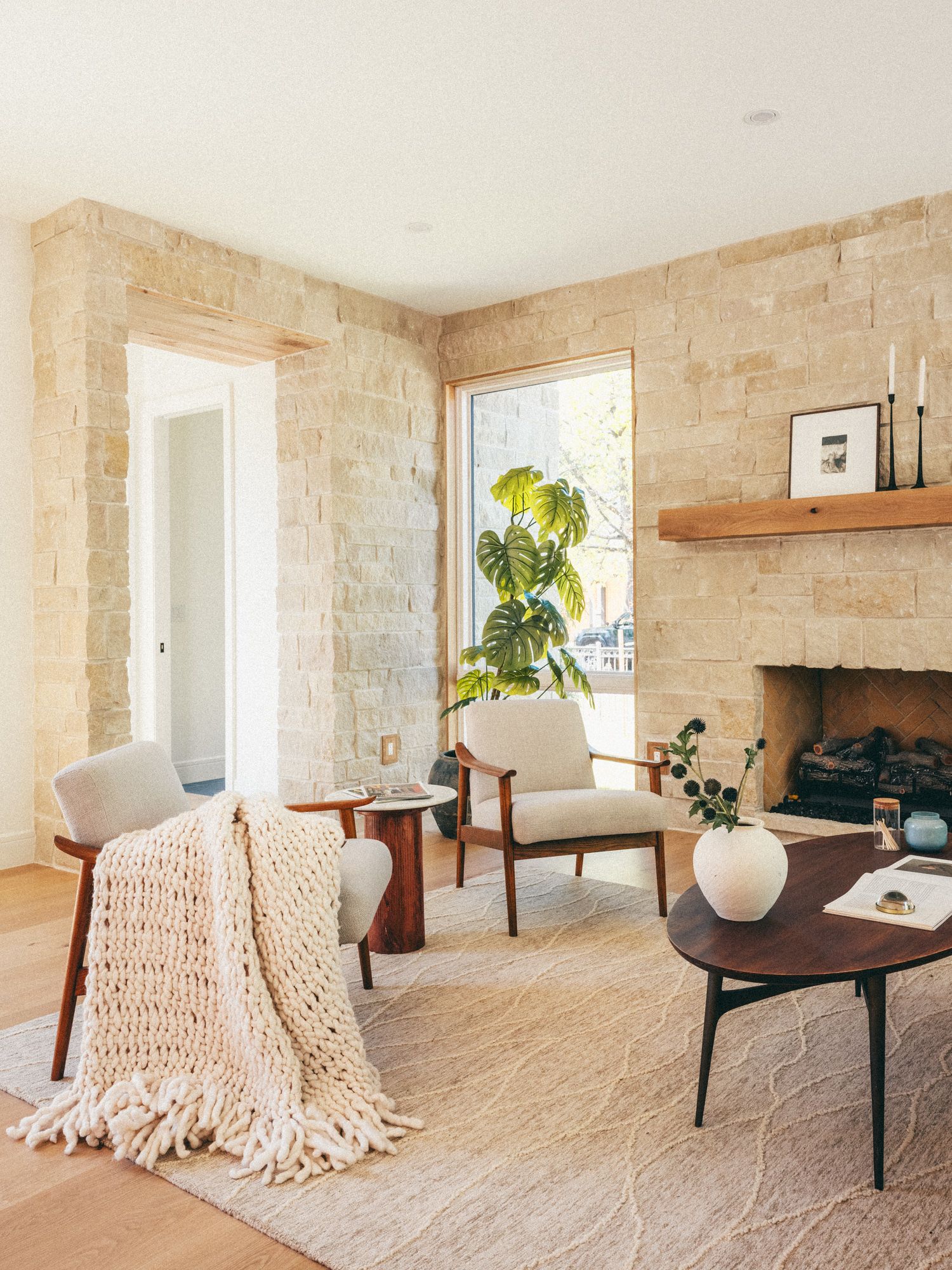 Warm living room with limestone walls, fireplace, mid-century chairs, chunky knit throw, and monstera plant on hardwood floors.