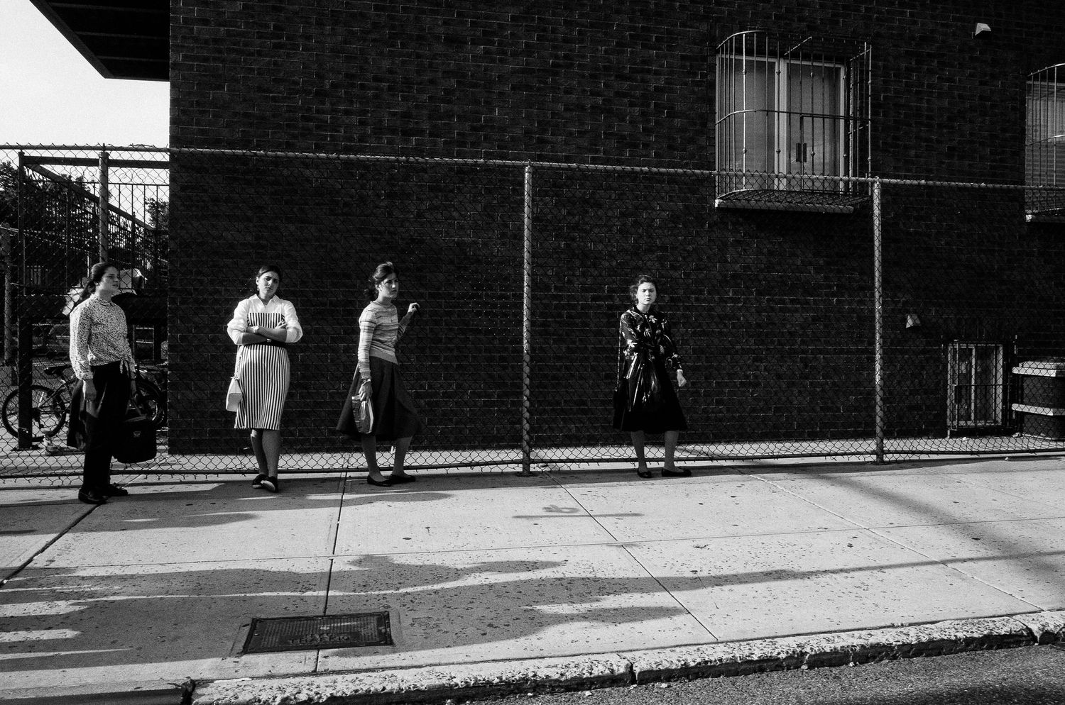 A black and white street scene shows people walking along a brick building wall with a chain link fence in an urban setting.