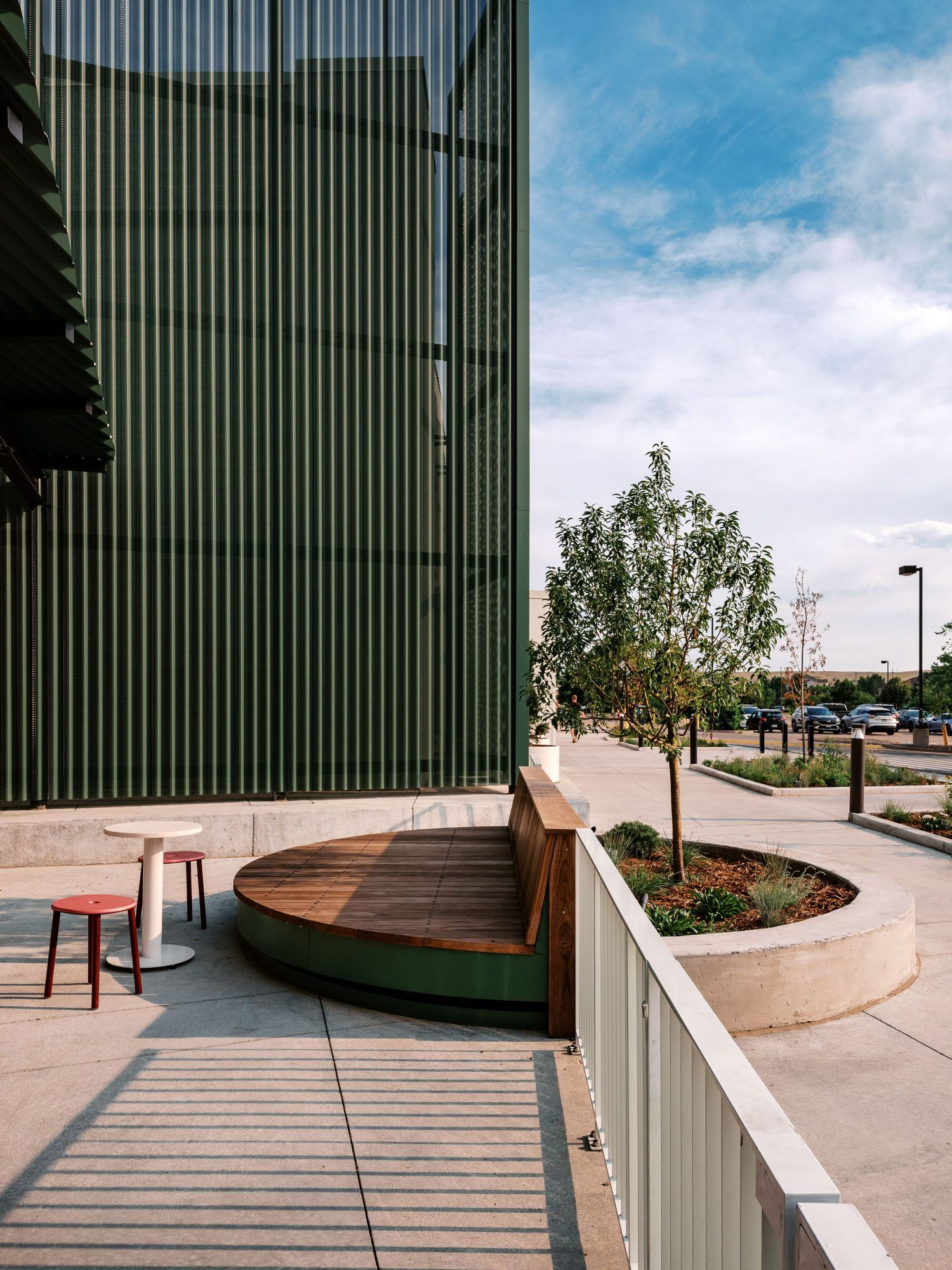 Curved wooden benches and planters line a modern concrete walkway against a green metal wall.