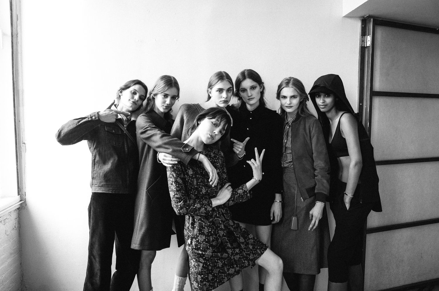 Black and white photo of eight young women posing together in stylish dark outfits in a bright room.