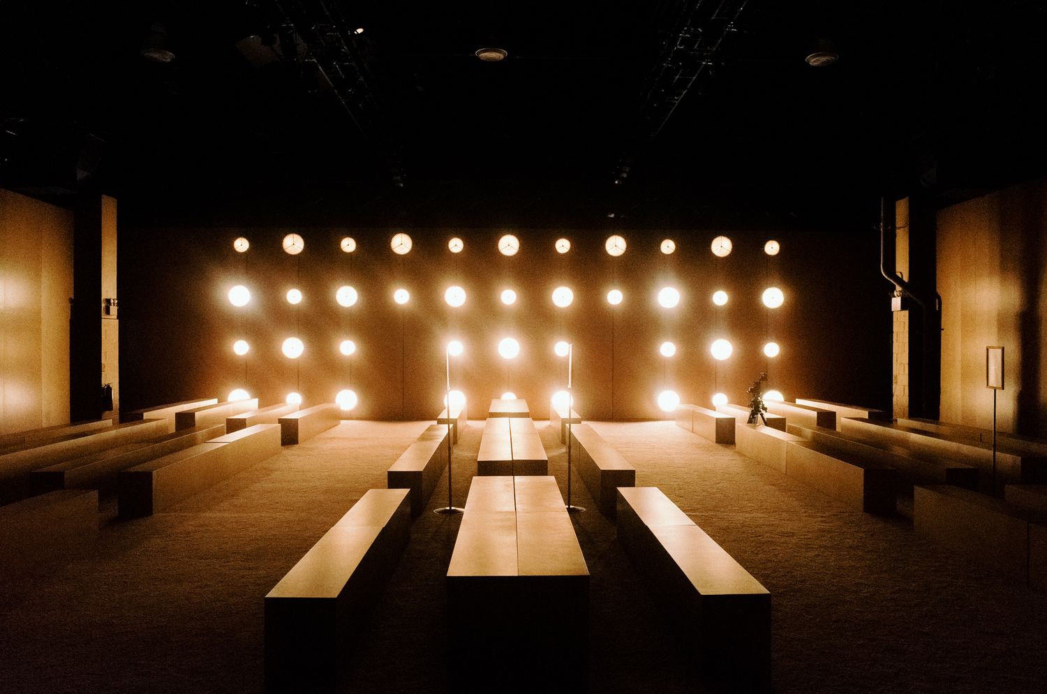 Minimalist event space with wooden benches, warm glowing round lights on back wall, dark ambiance.