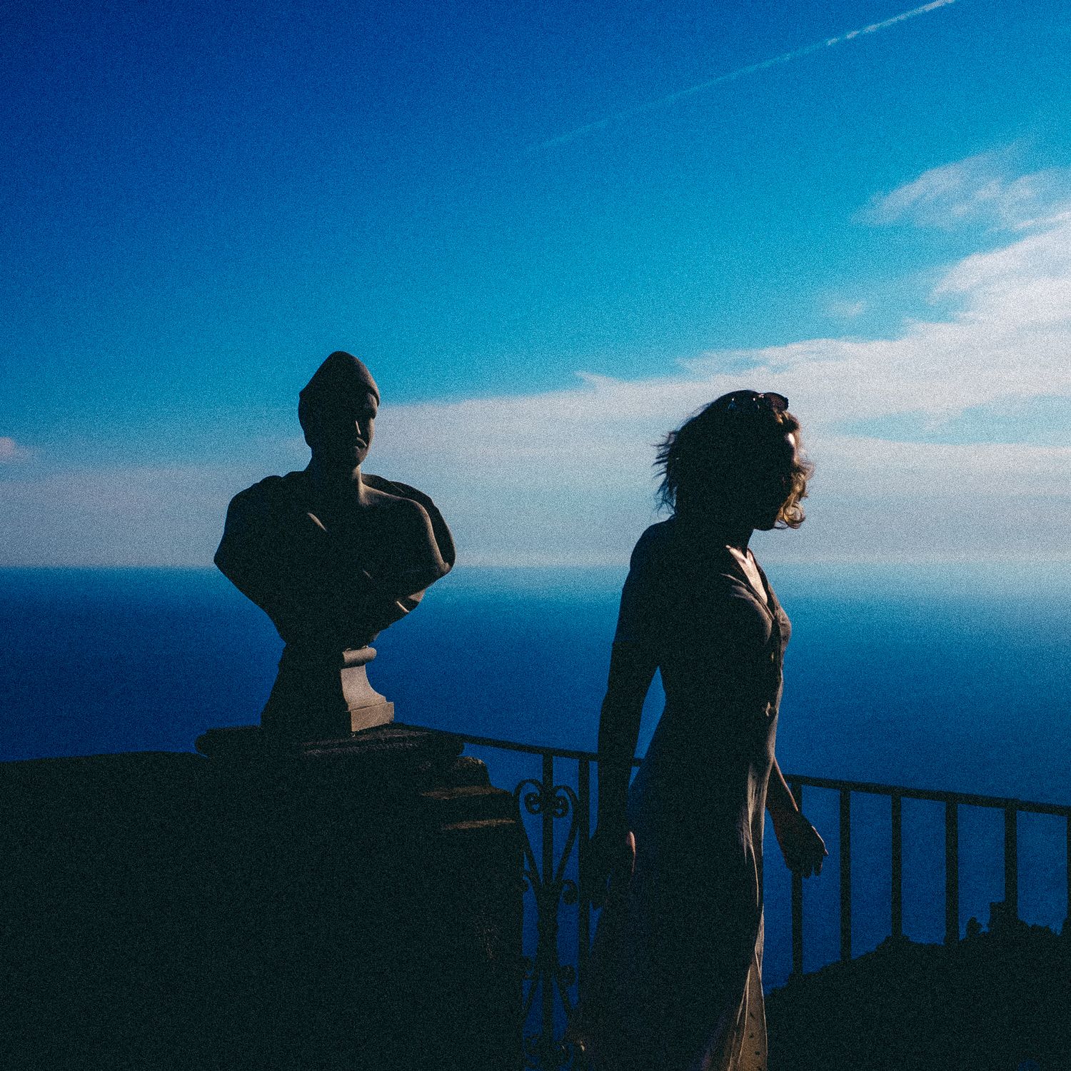 Silhouetted figures stand against a vibrant blue ocean horizon at a scenic coastal viewpoint during sunset.
