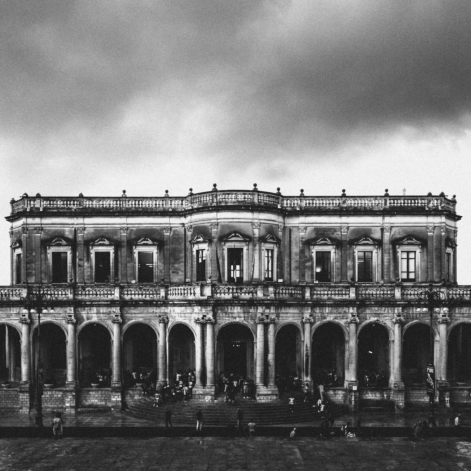 A black and white panoramic photograph of a historic colonnade building with arched walkways and uniform architecture.