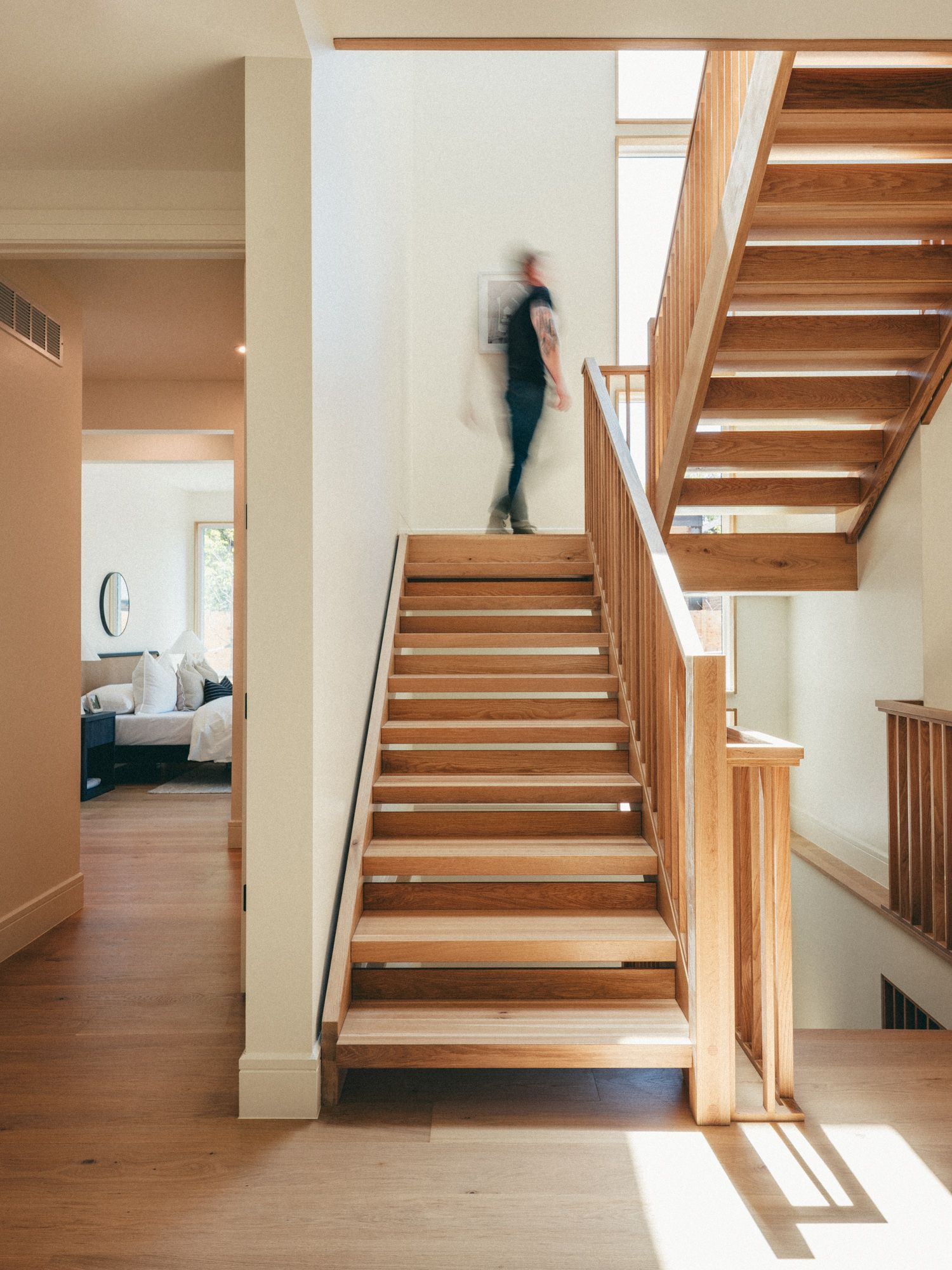 Open wooden staircase with light oak steps, glass railings, and a blurred figure ascending in a bright home.