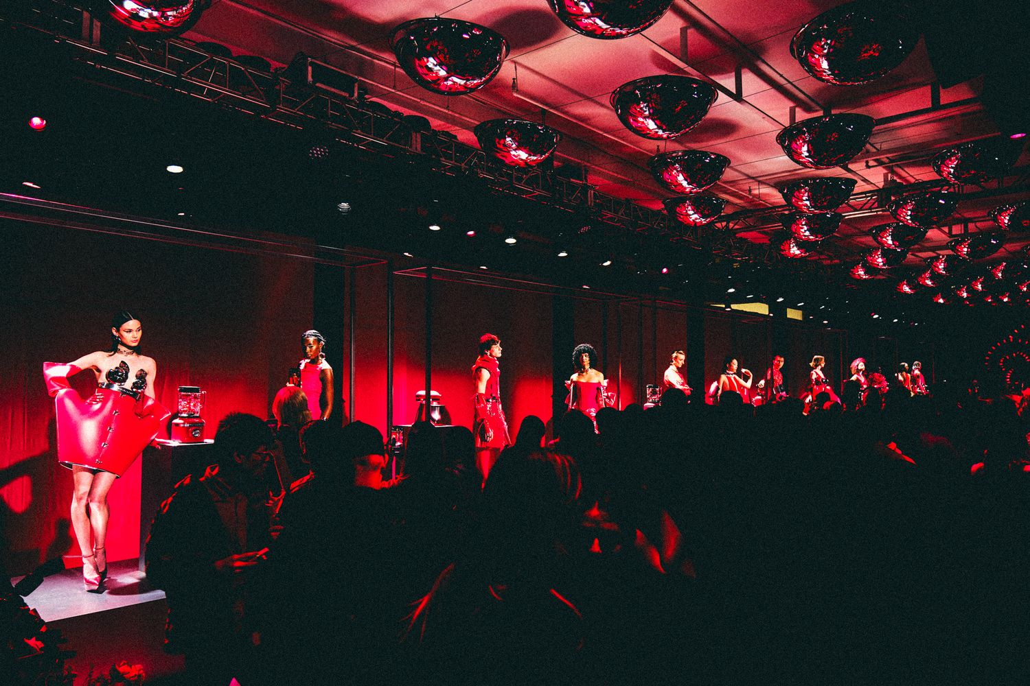 Performers in red costumes on a dimly lit stage with mirror ball ceiling lights and a seated audience.