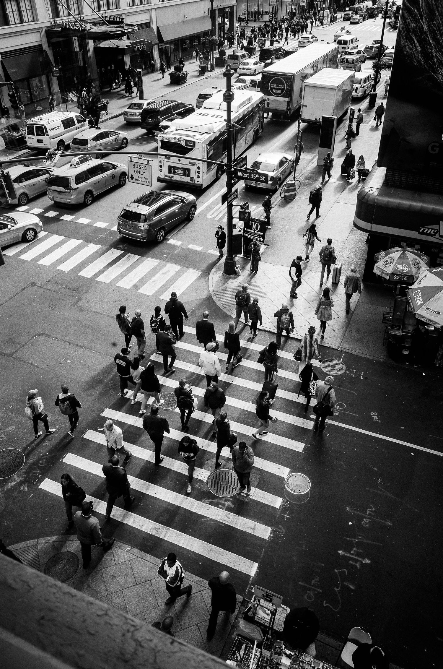 Black and white aerial view of pedestrians crossing a busy intersection in a city with cars and buses on the street.