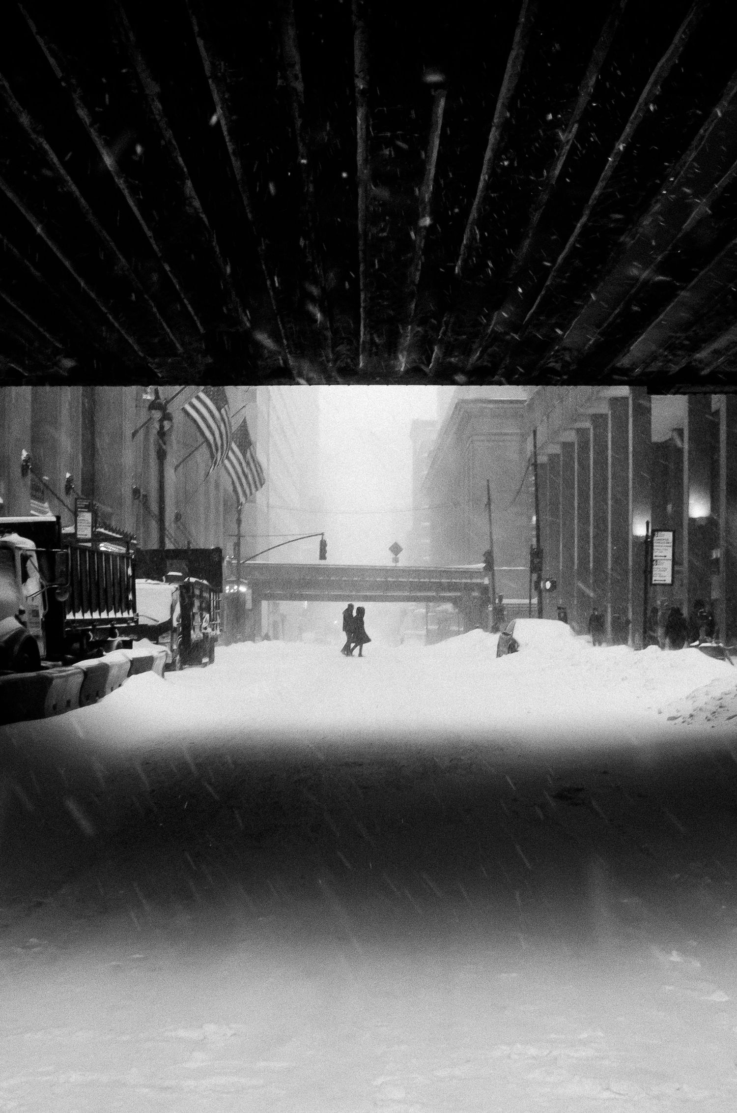 A black and white panoramic view of a snowy street at night with illuminated buildings and streetlights casting shadows.