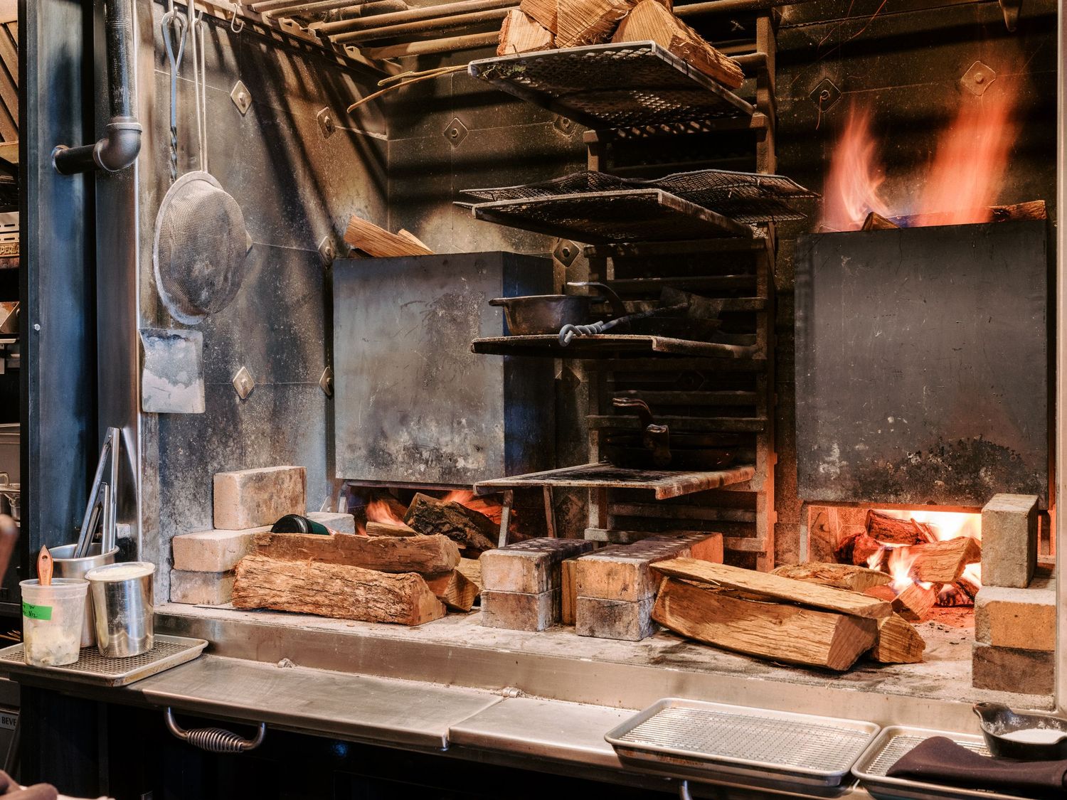 Professional kitchen with stainless steel equipment and wood-fired cooking station.