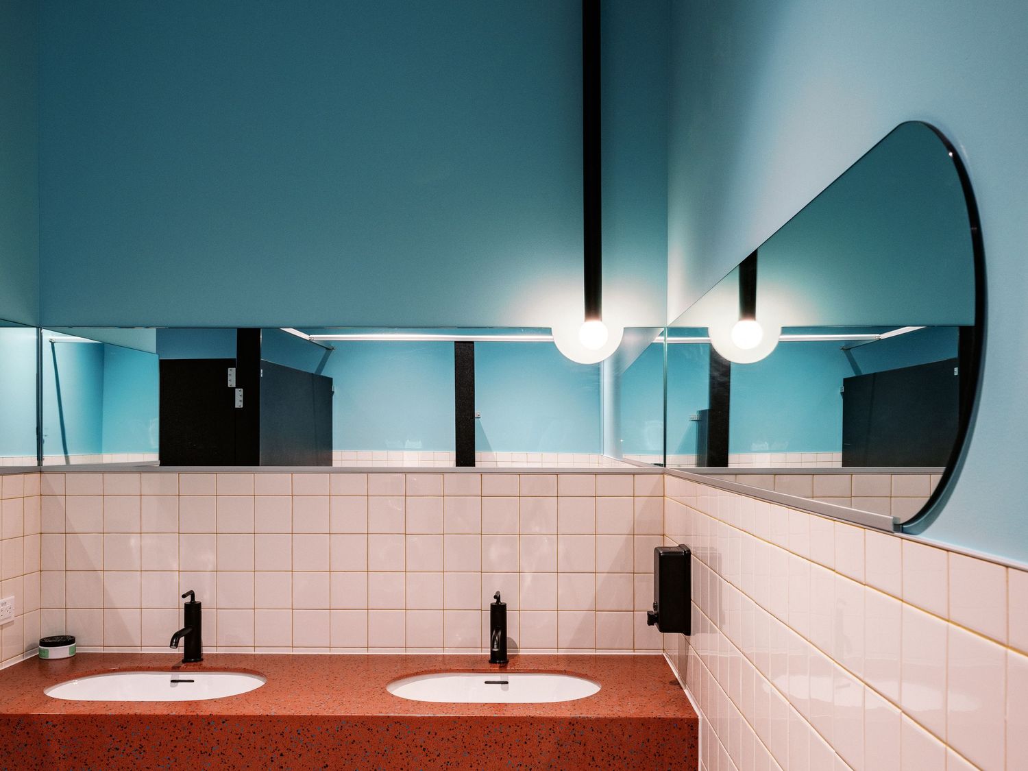 Modern bathroom interior with round mirror, white tile walls, and dual sinks on coral countertop.