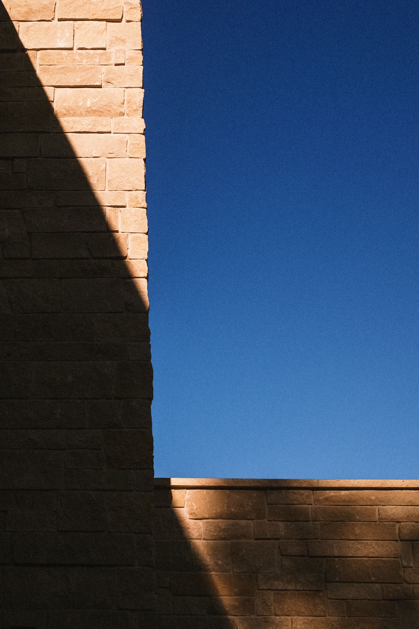 Close-up of sunlit limestone brick wall casting dramatic shadow against a clear deep blue sky.
