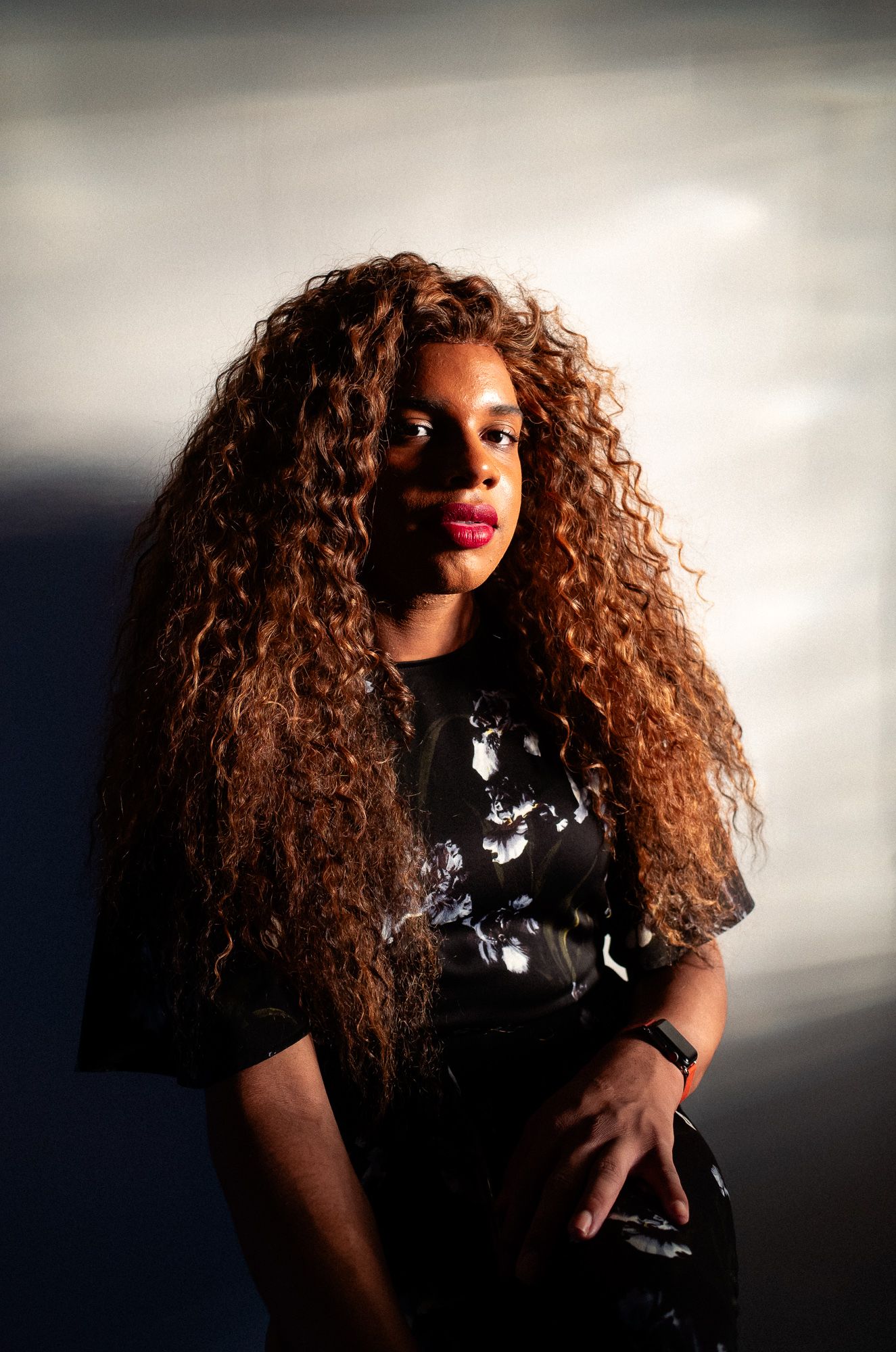 Portrait of someone with long curly auburn hair wearing a dark patterned outfit against moody lighting.