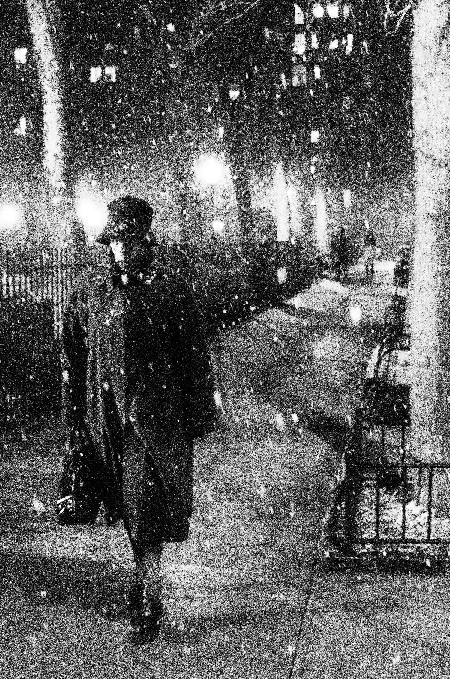 Black and white photograph of snowy city street at night with silhouettes of people and glowing streetlights.
