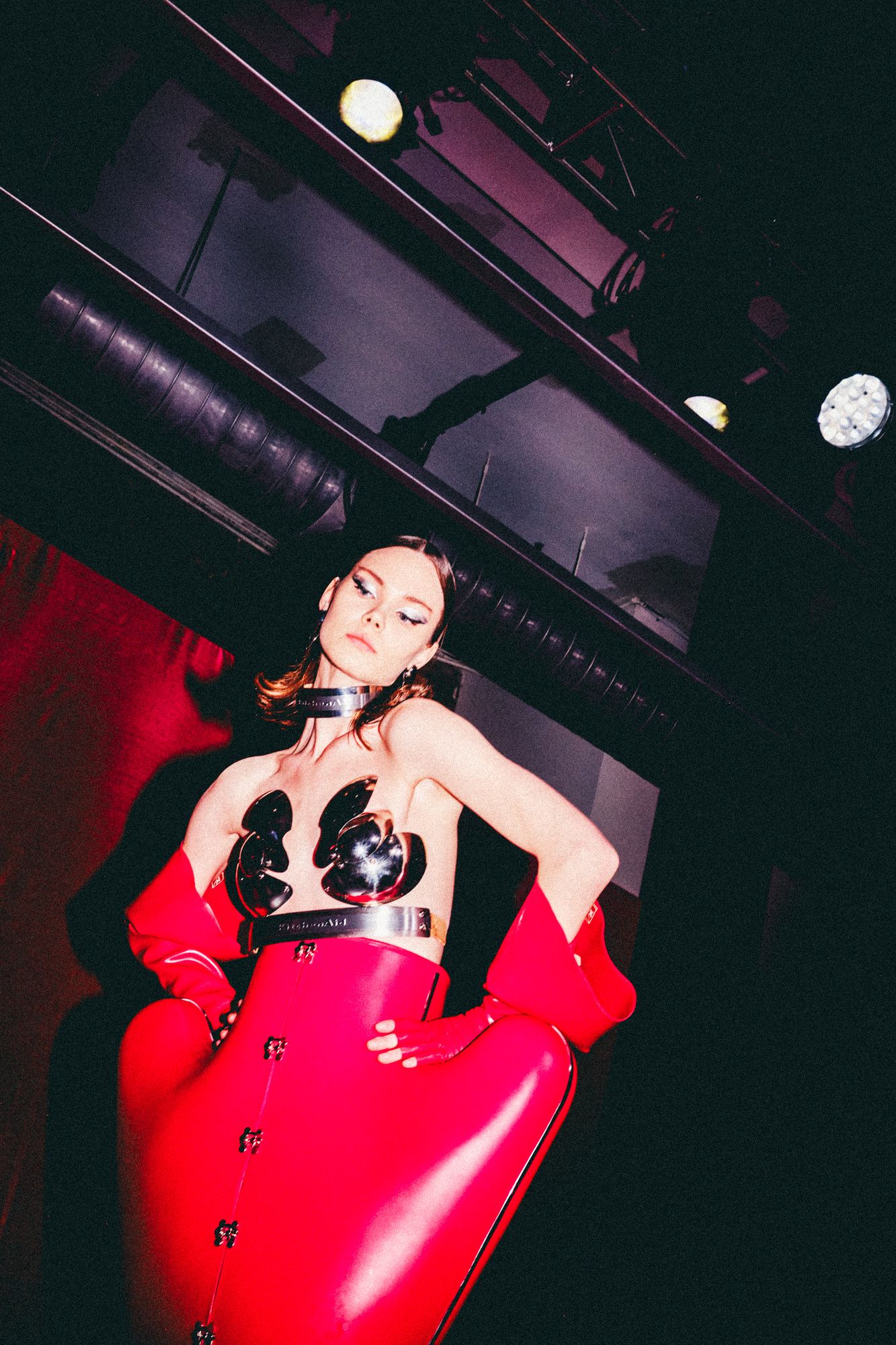 Woman in red latex coat and black bustier poses dramatically under stage lights in a dark venue.