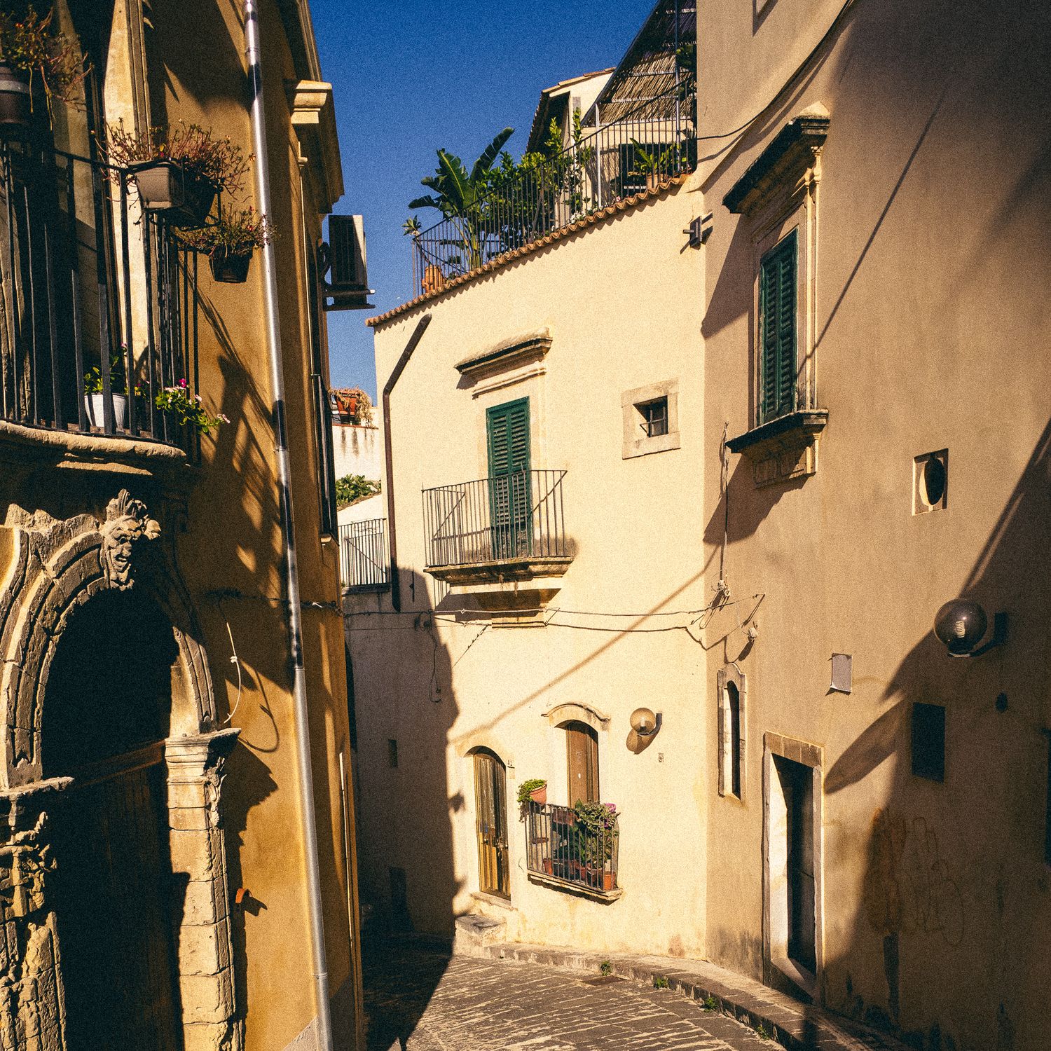 Narrow cobblestone street with traditional cream-colored buildings and balconies under bright blue sky in Mediterranean town.