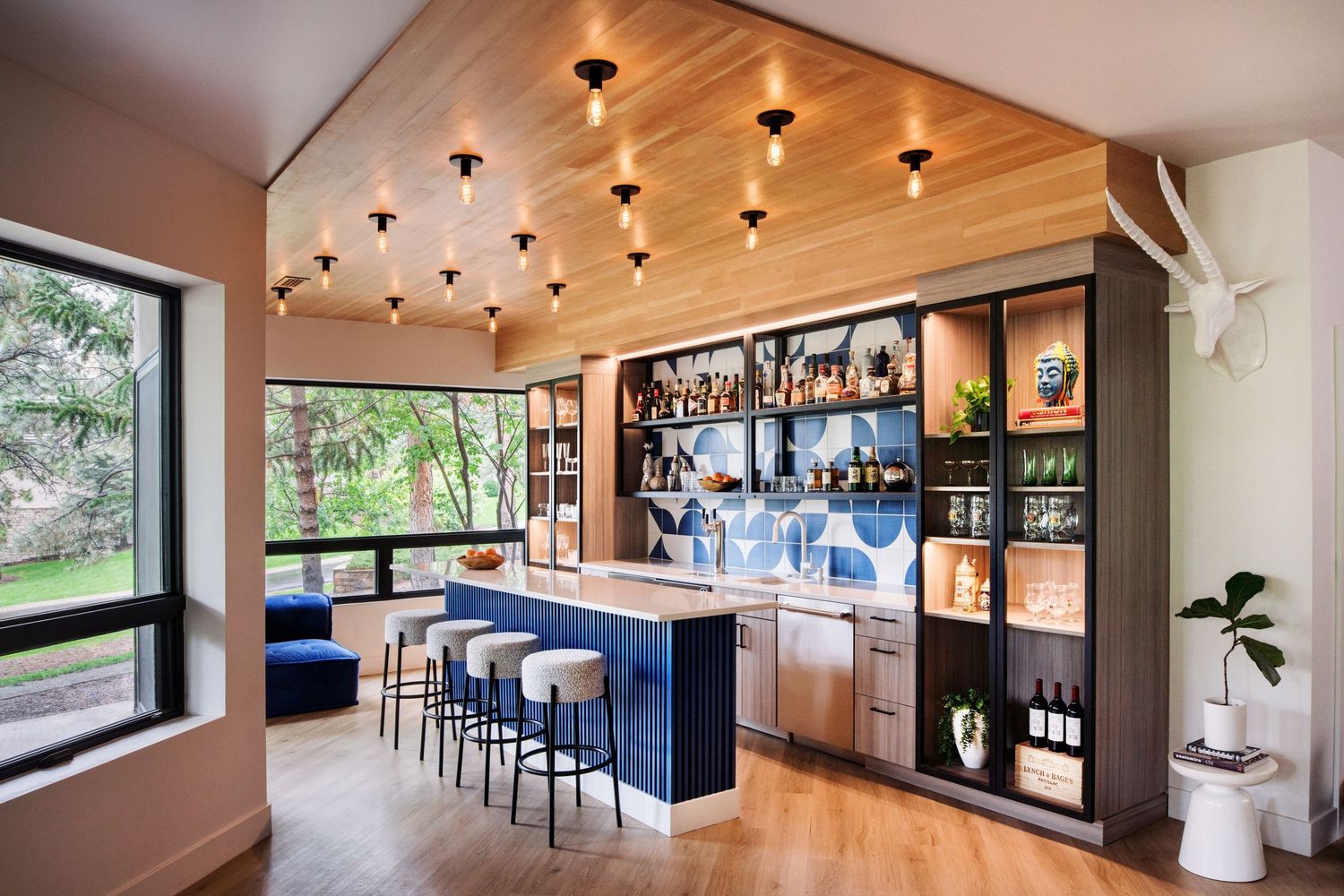Modern home bar with wood ceiling, pendant lights, and large window overlooking nature.