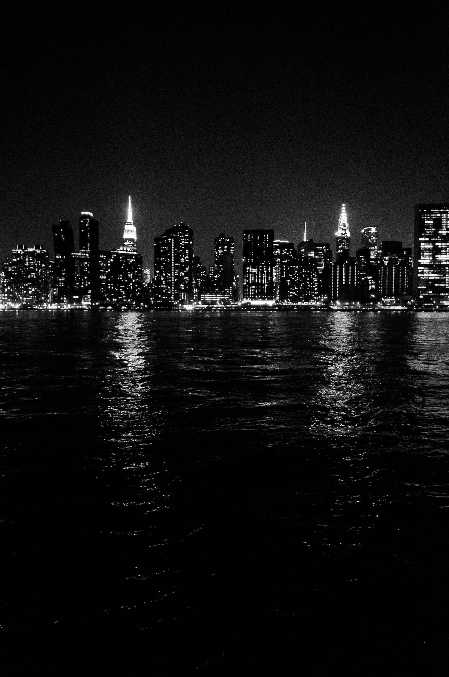 Black and white nighttime skyline of Manhattan with illuminated skyscrapers reflecting on the East River.