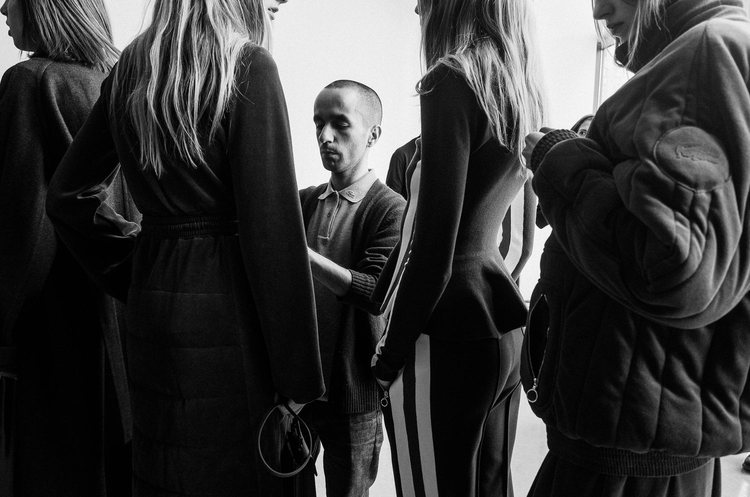 A black and white photograph shows people standing in a group having a conversation in a minimalist setting.