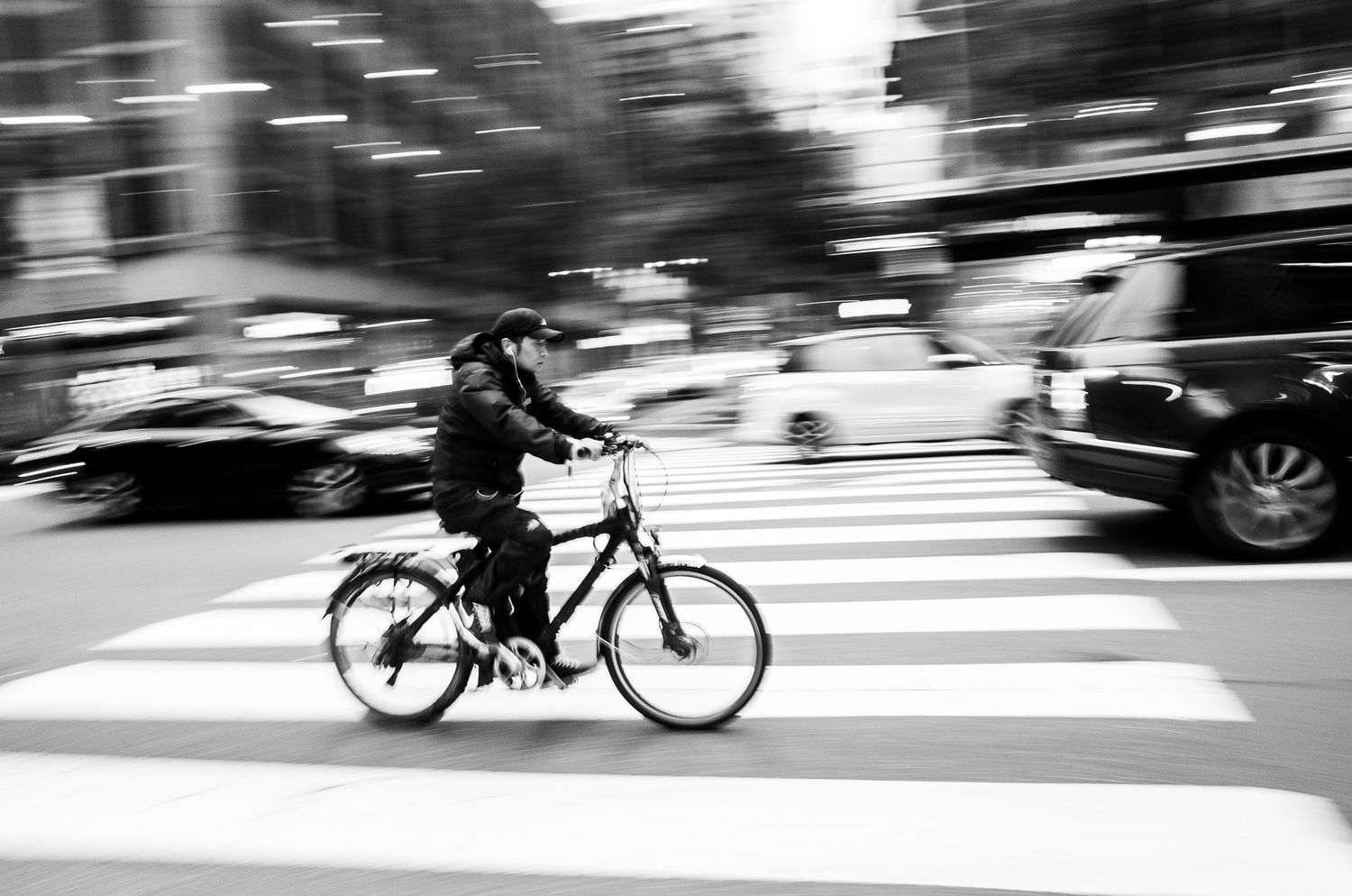 A cyclist in motion crosses a busy city street on a bicycle, captured in black and white with motion blur effect.