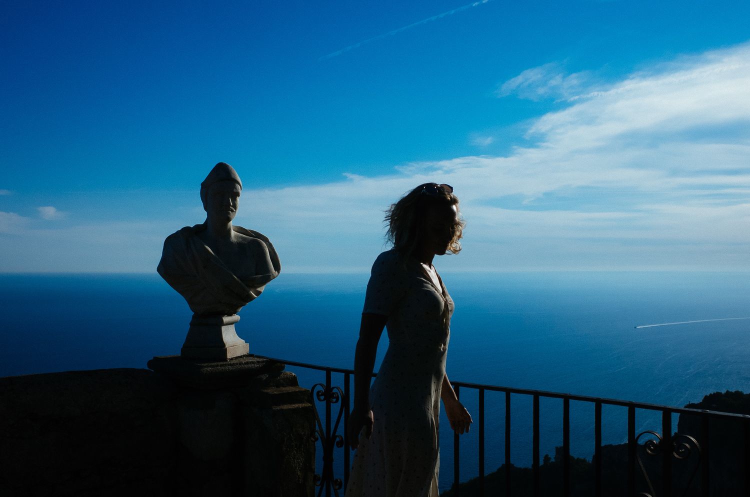 Silhouettes of people against bright blue sky and ocean view from elevated viewpoint.