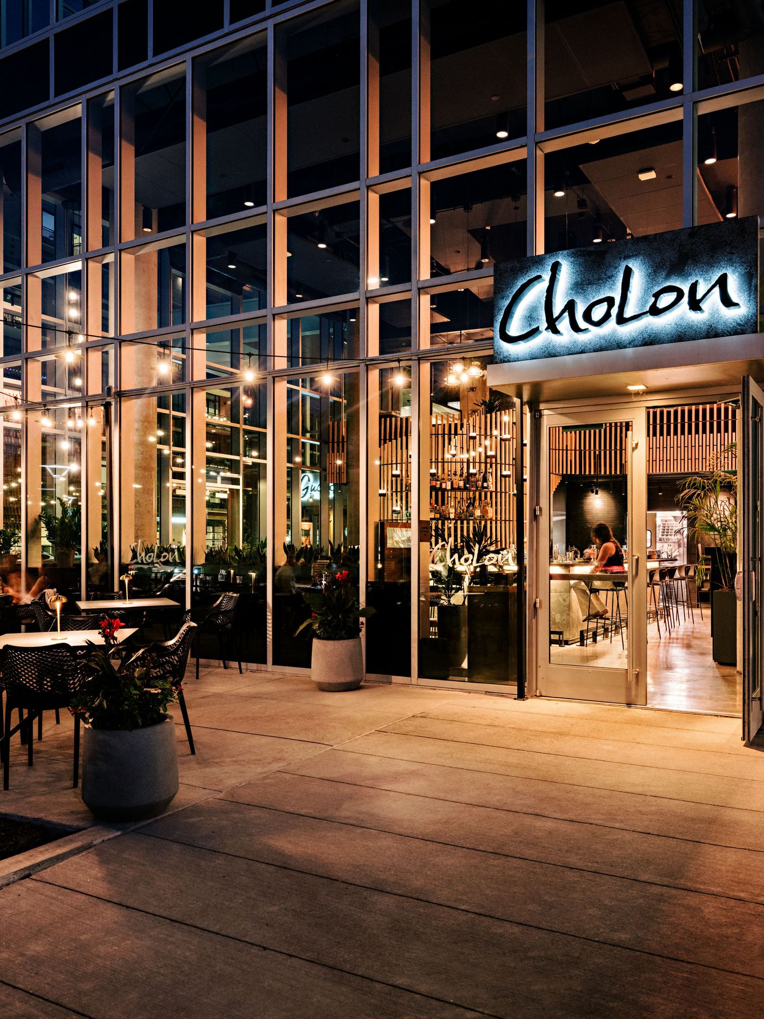 Evening exterior view of illuminated restaurant storefront with glass walls and outdoor seating.