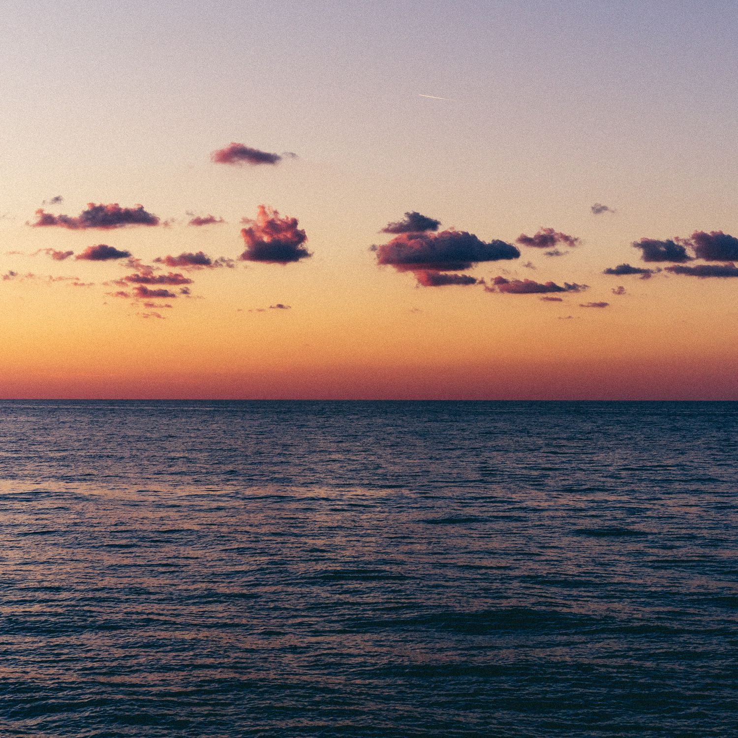 Panoramic view of a tranquil ocean horizon at sunset with scattered clouds reflecting warm orange and purple hues.