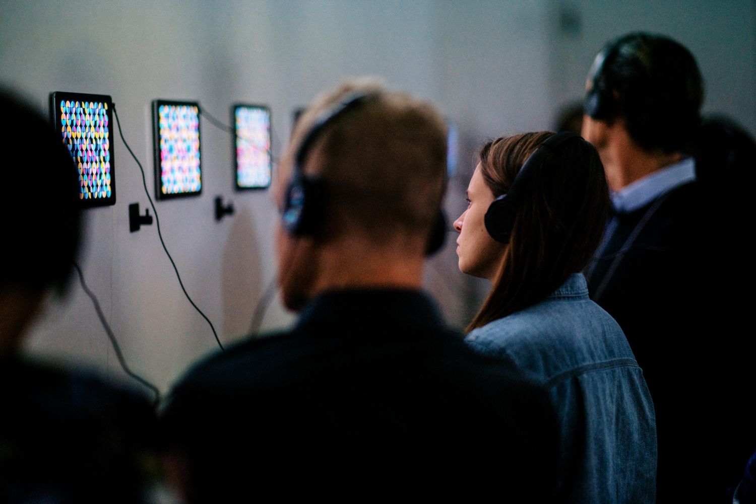 People wearing headphones viewing colorful digital art displays on tablets mounted on a gallery wall.