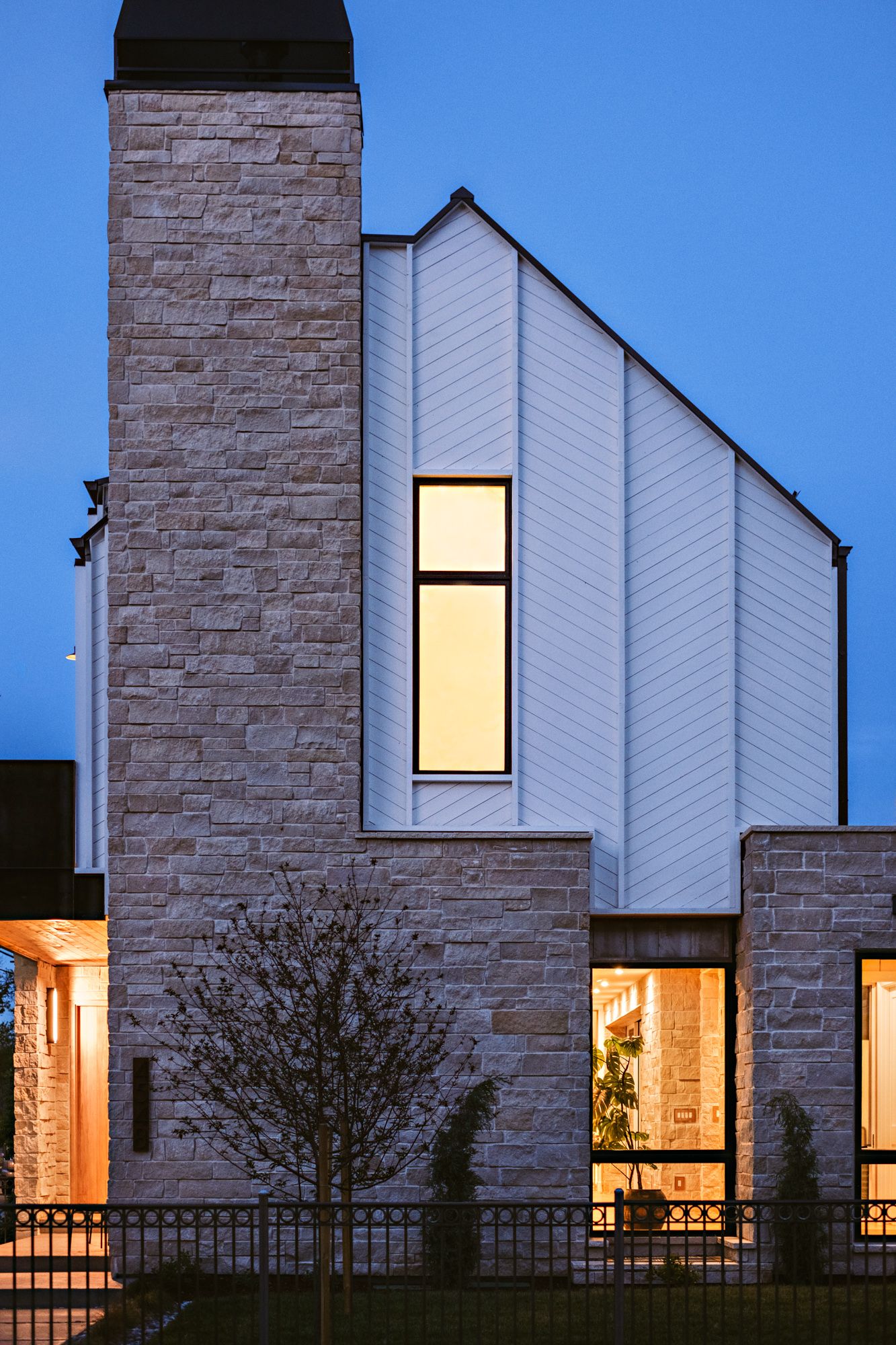 Modern home exterior at dusk featuring stone chimney, white vertical siding, and illuminated narrow windows.