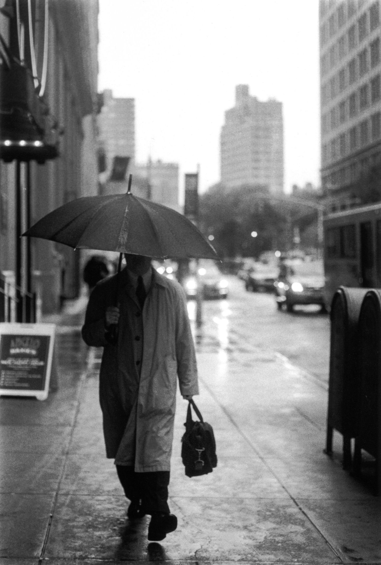 A black and white photograph shows a person walking with an umbrella on a rainy city street.